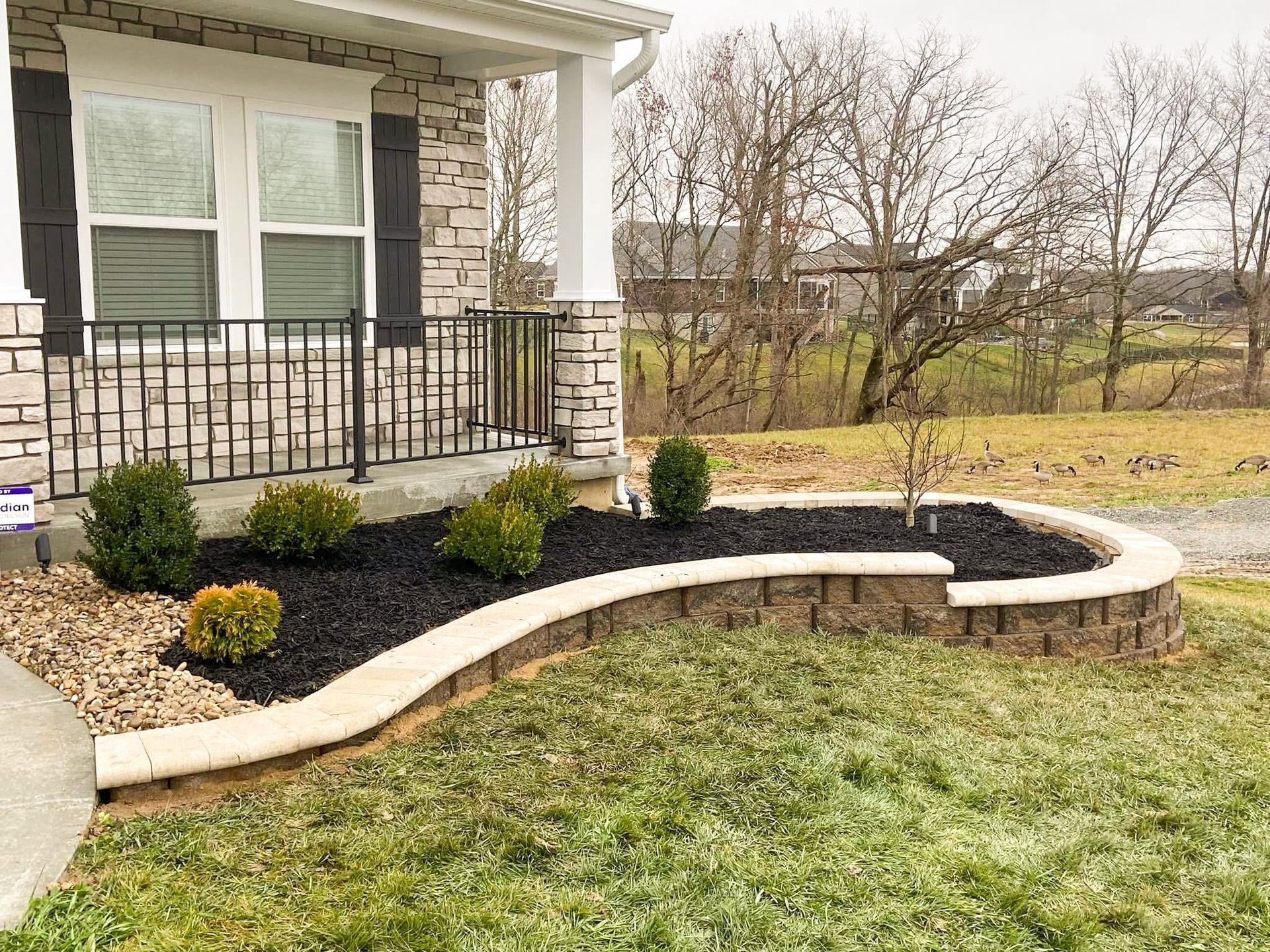 Curved stone retaining wall with black mulch, green shrubs, and a house with a porch.