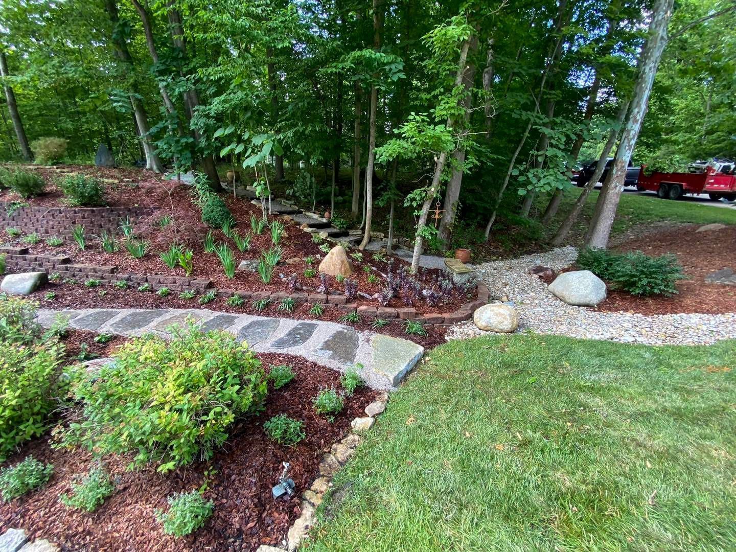 Landscaped yard with a stone path, mulch beds, and a small stream leading into a wooded area.