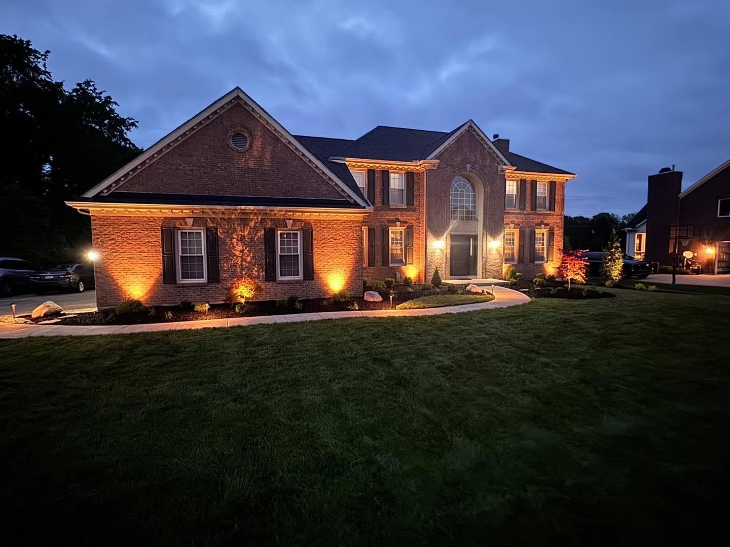Brick house illuminated with warm spotlights at dusk. Landscaping and lawn in the foreground.
