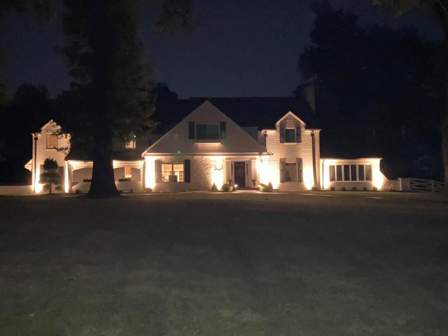 Large, lit-up house at night with floodlights illuminating the facade, set on a dark lawn.