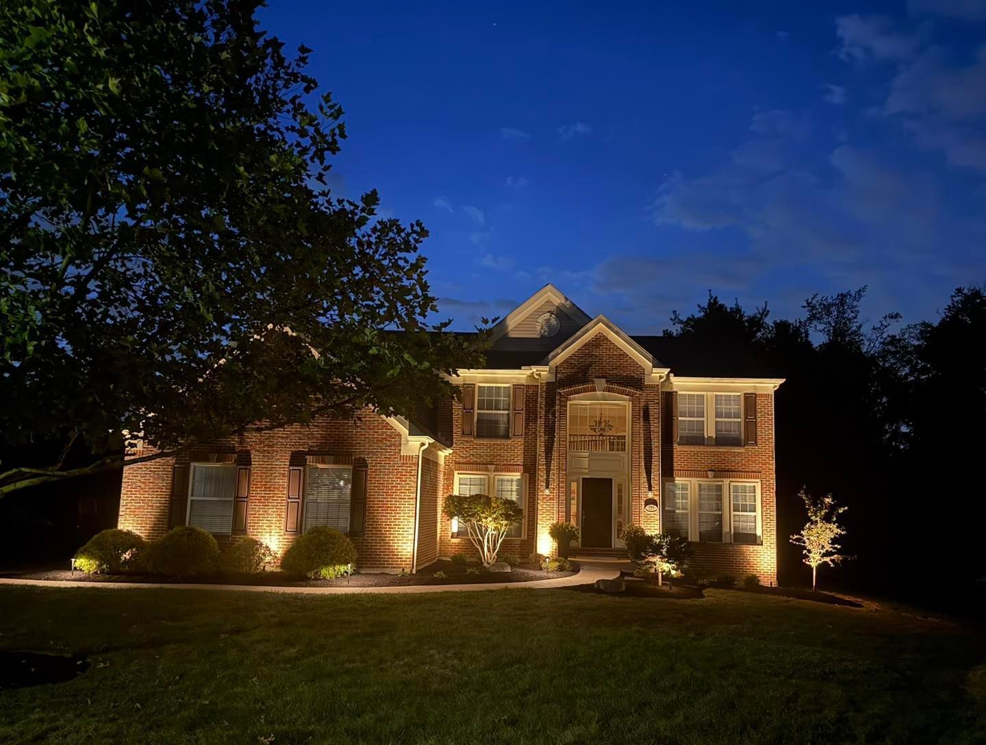Brick house illuminated at night with landscape lighting. Green lawn and dark blue sky.