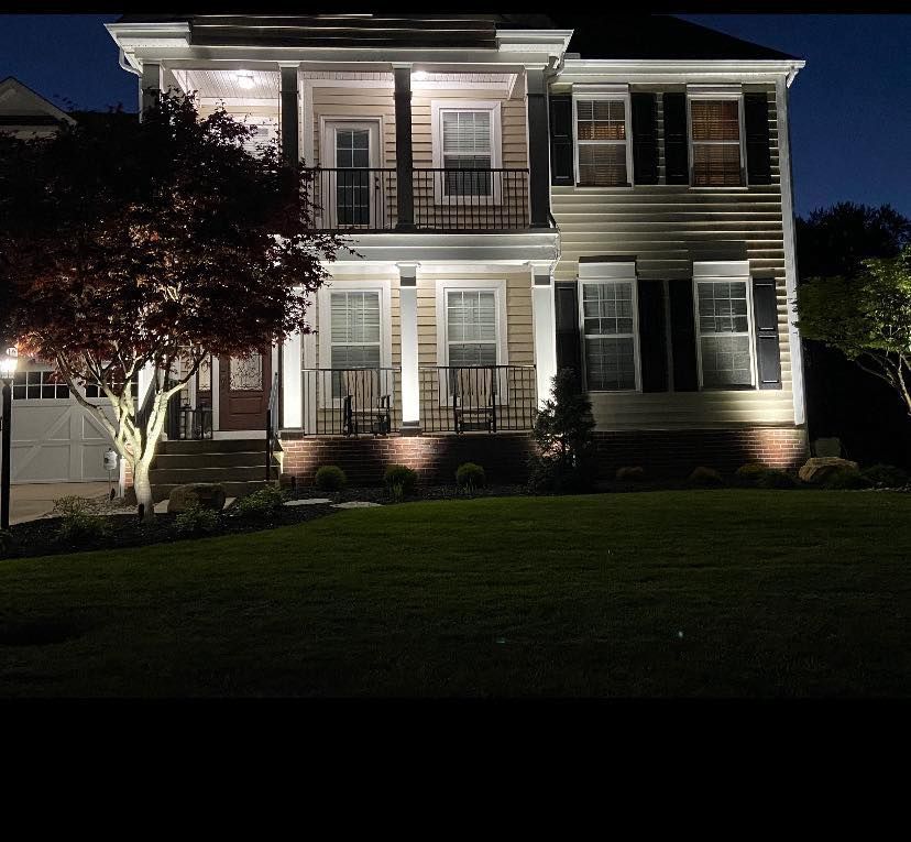 Nighttime view of a two-story house with outdoor lighting illuminating the facade, front porch, and landscaping.