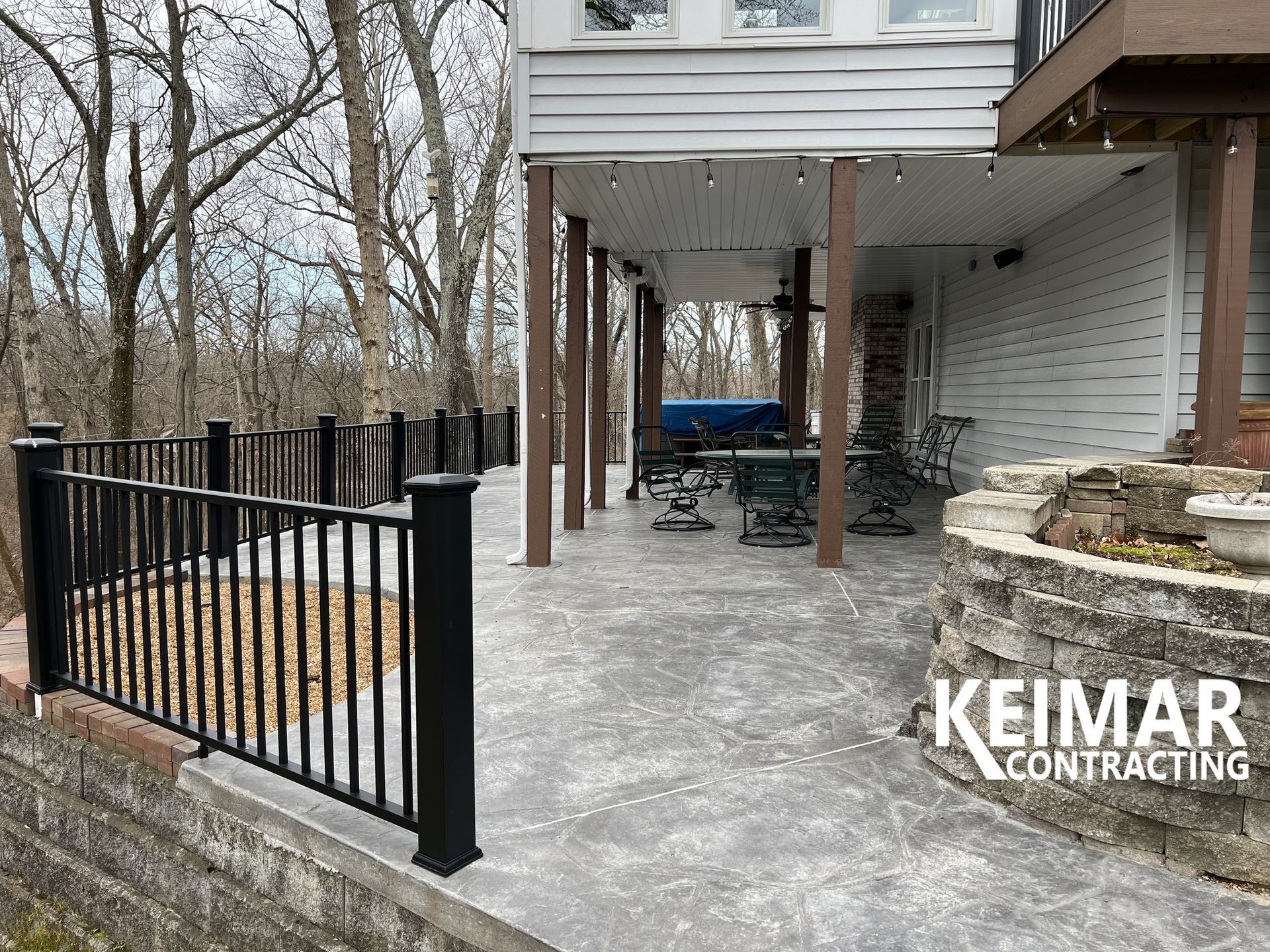 Black railing and deck overlooking a patio area. House with brown columns and trees in the background.