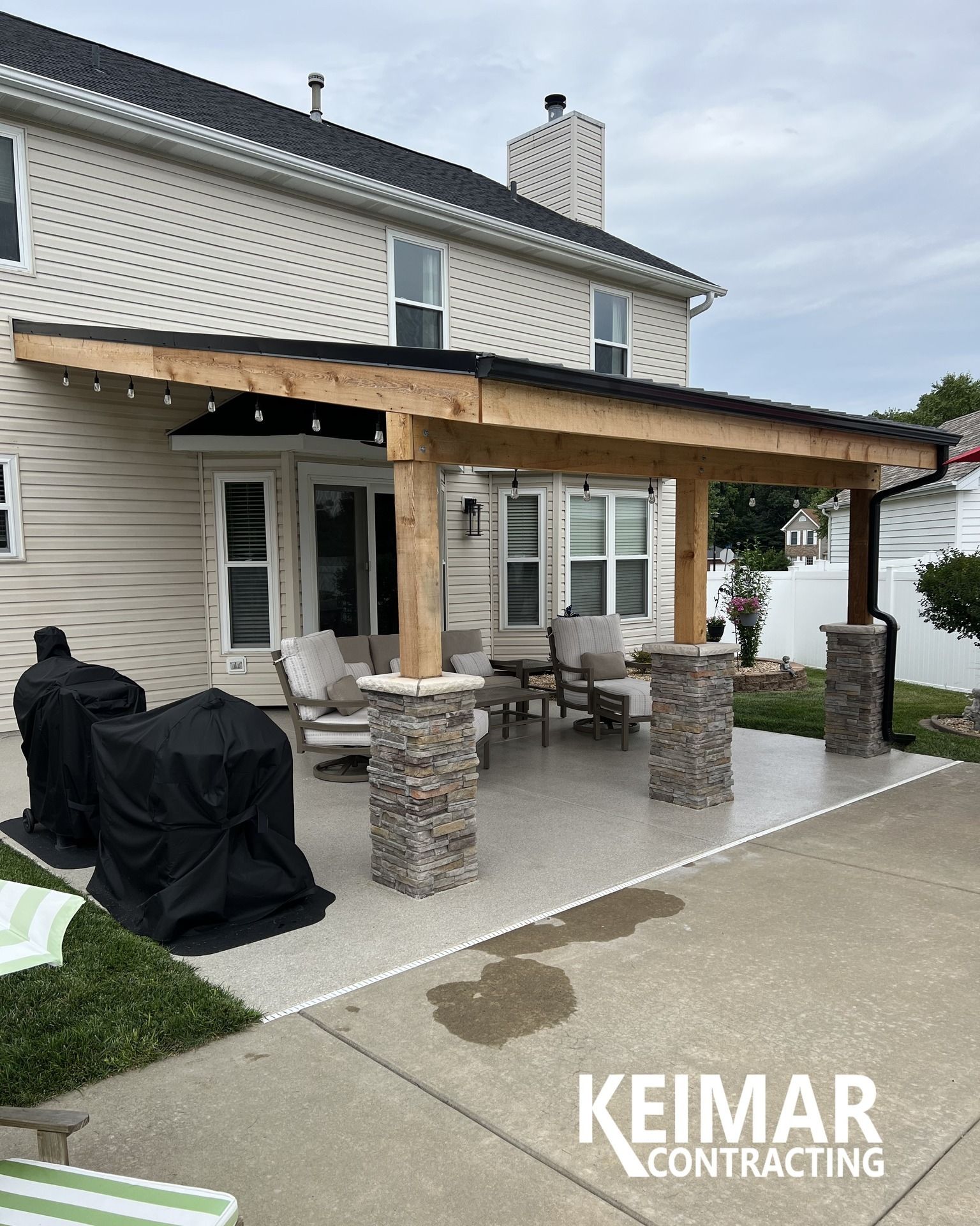 Patio with stone columns, concrete floor, wooden pergola, and outdoor furniture next to a two-story house.