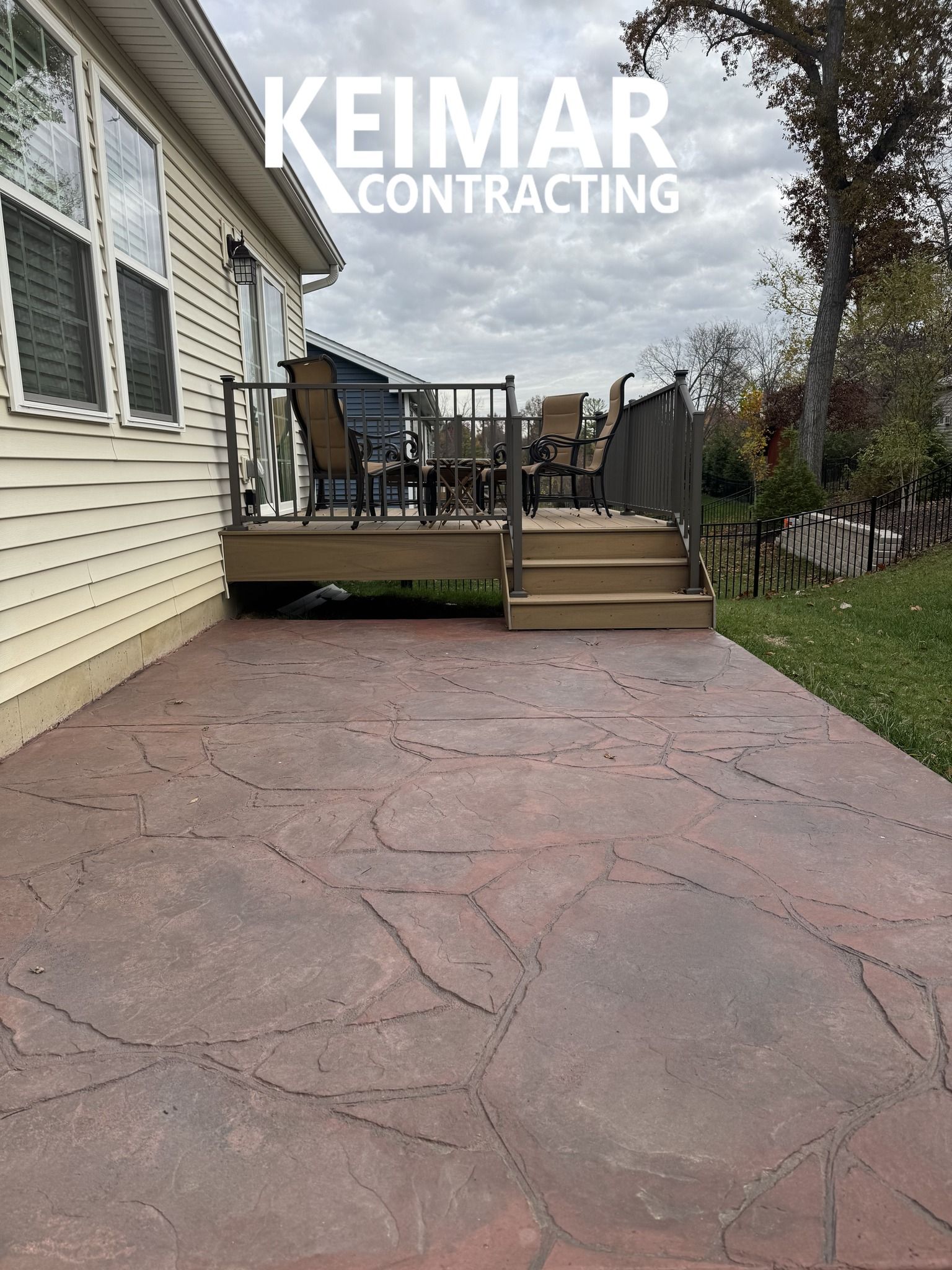 Stamped concrete patio leading to a wooden deck with railing; overcast sky.