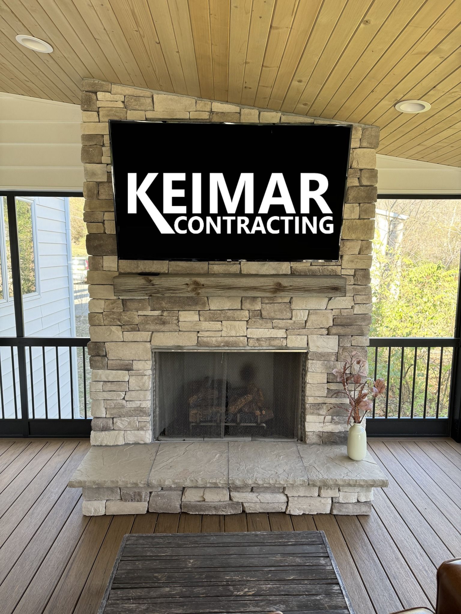 Stone fireplace with Keimar Contracting logo, wooden ceiling, and deck.