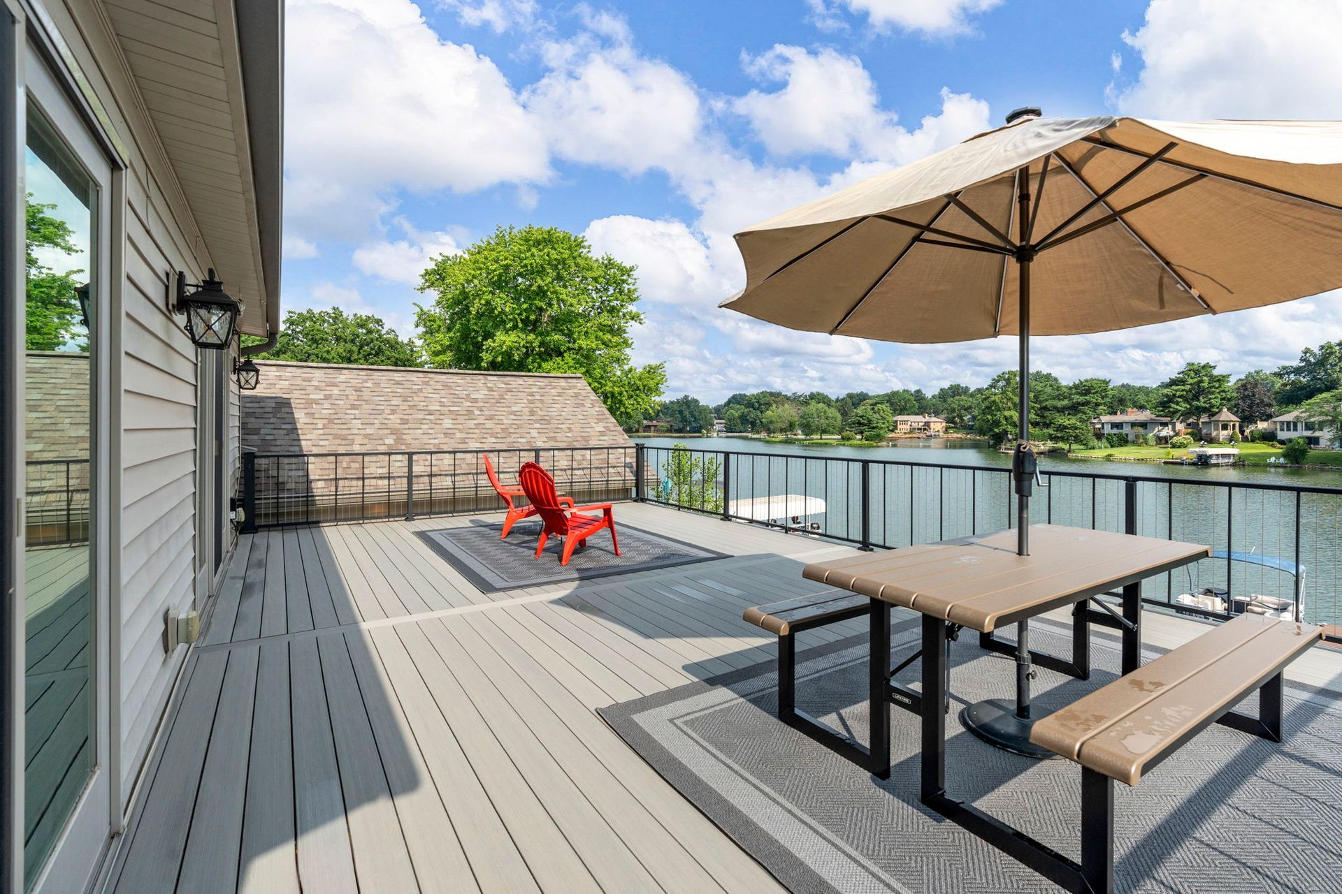 Deck overlooking a lake, with a picnic table, umbrella, and red chairs.