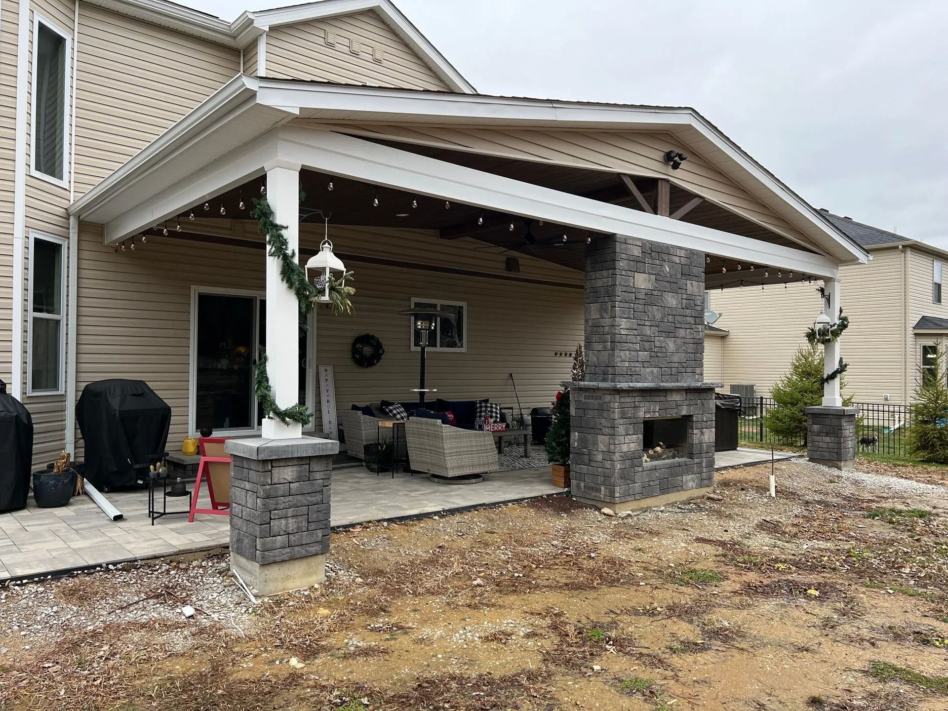 Covered outdoor patio with a stone fireplace and columns. Beige siding, string lights, and seating visible.