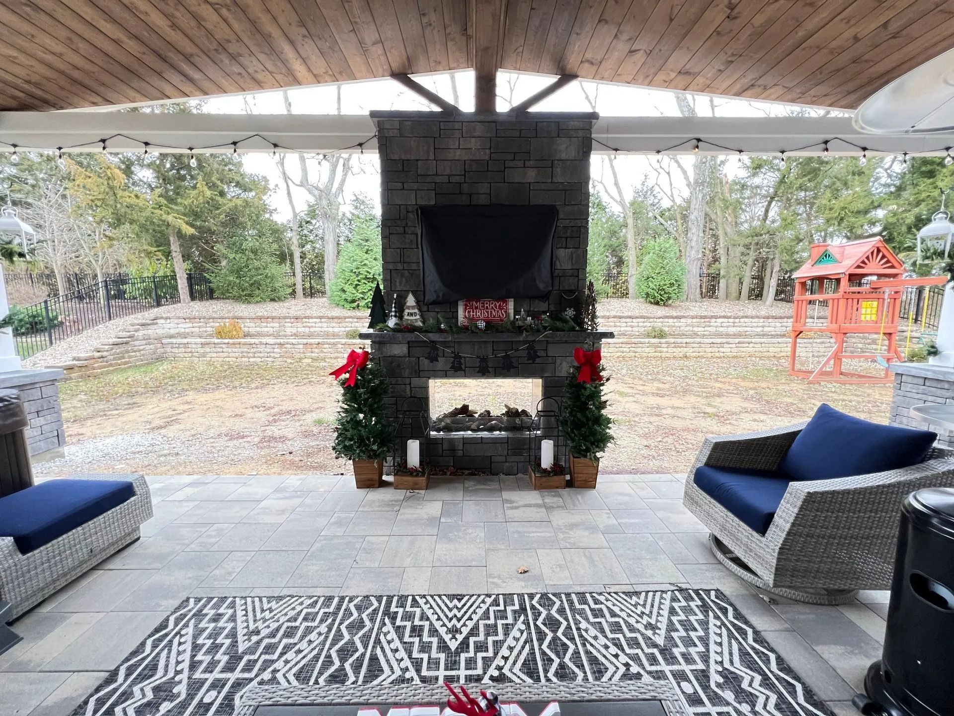 Patio with fireplace, seating, rug, and small Christmas trees, under a covered structure with a yard in the background.