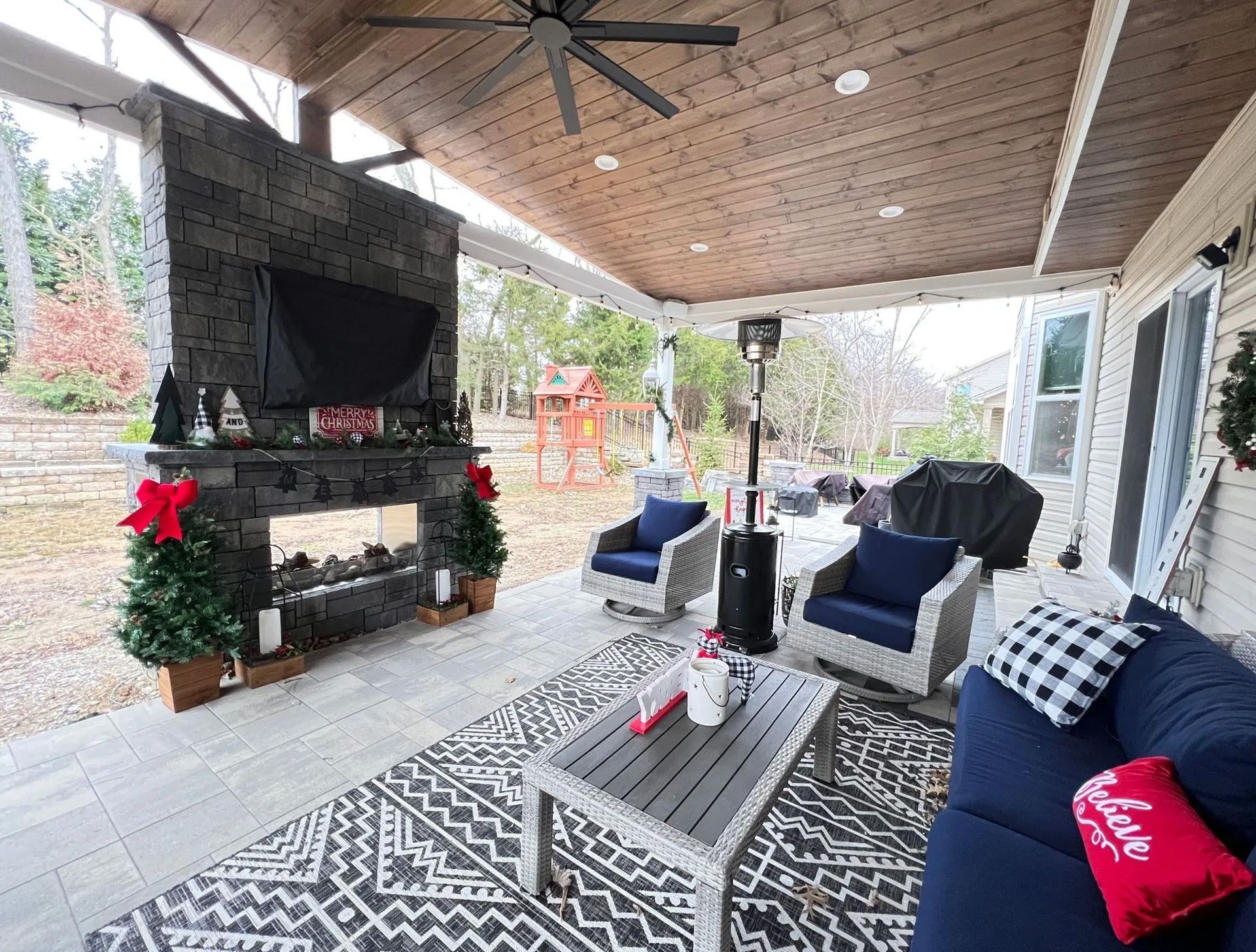 Covered patio with fireplace, seating, and holiday decorations, overlooking a yard.