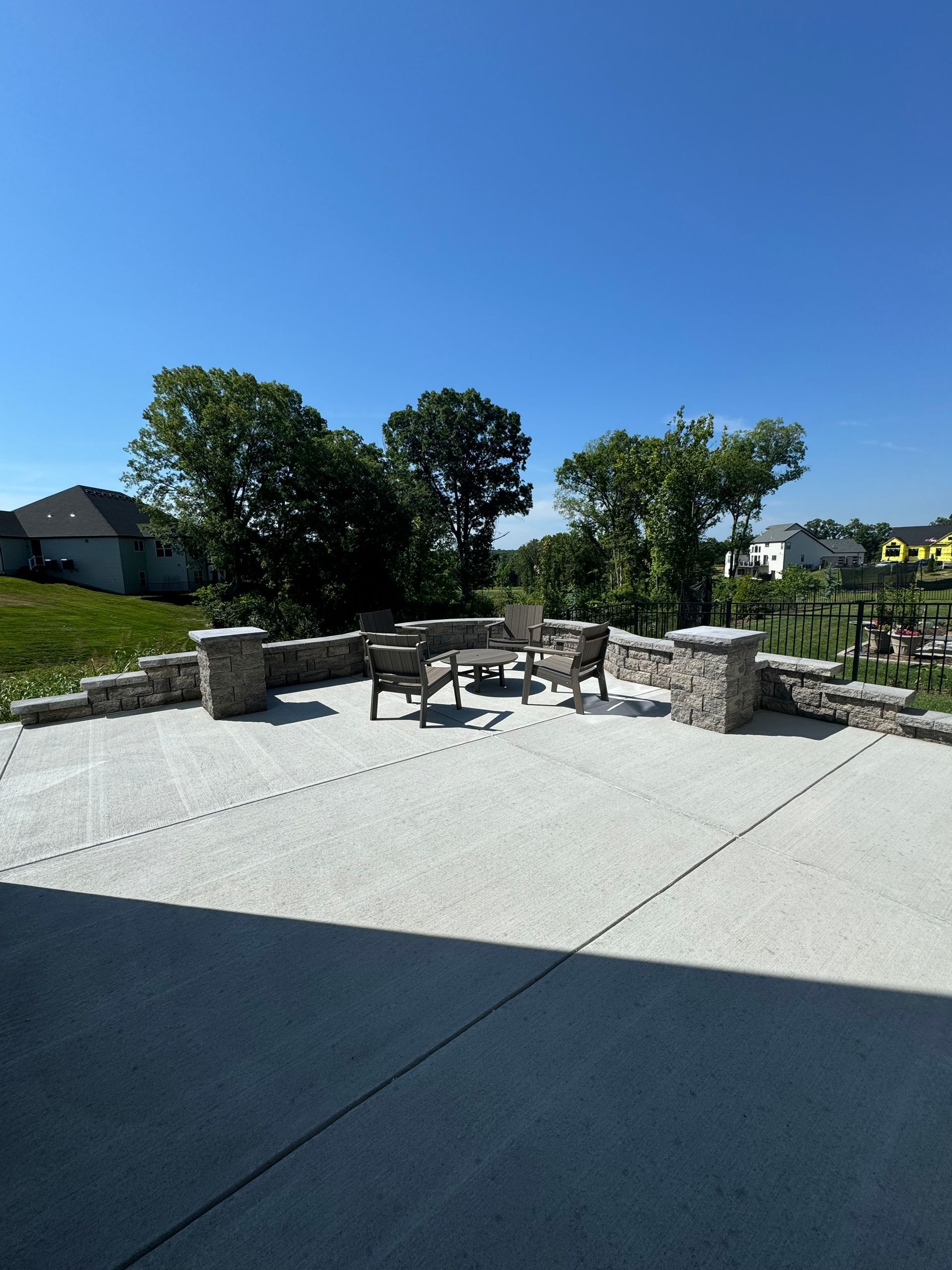 Outdoor patio with stone columns and seating, surrounded by trees under a blue sky.