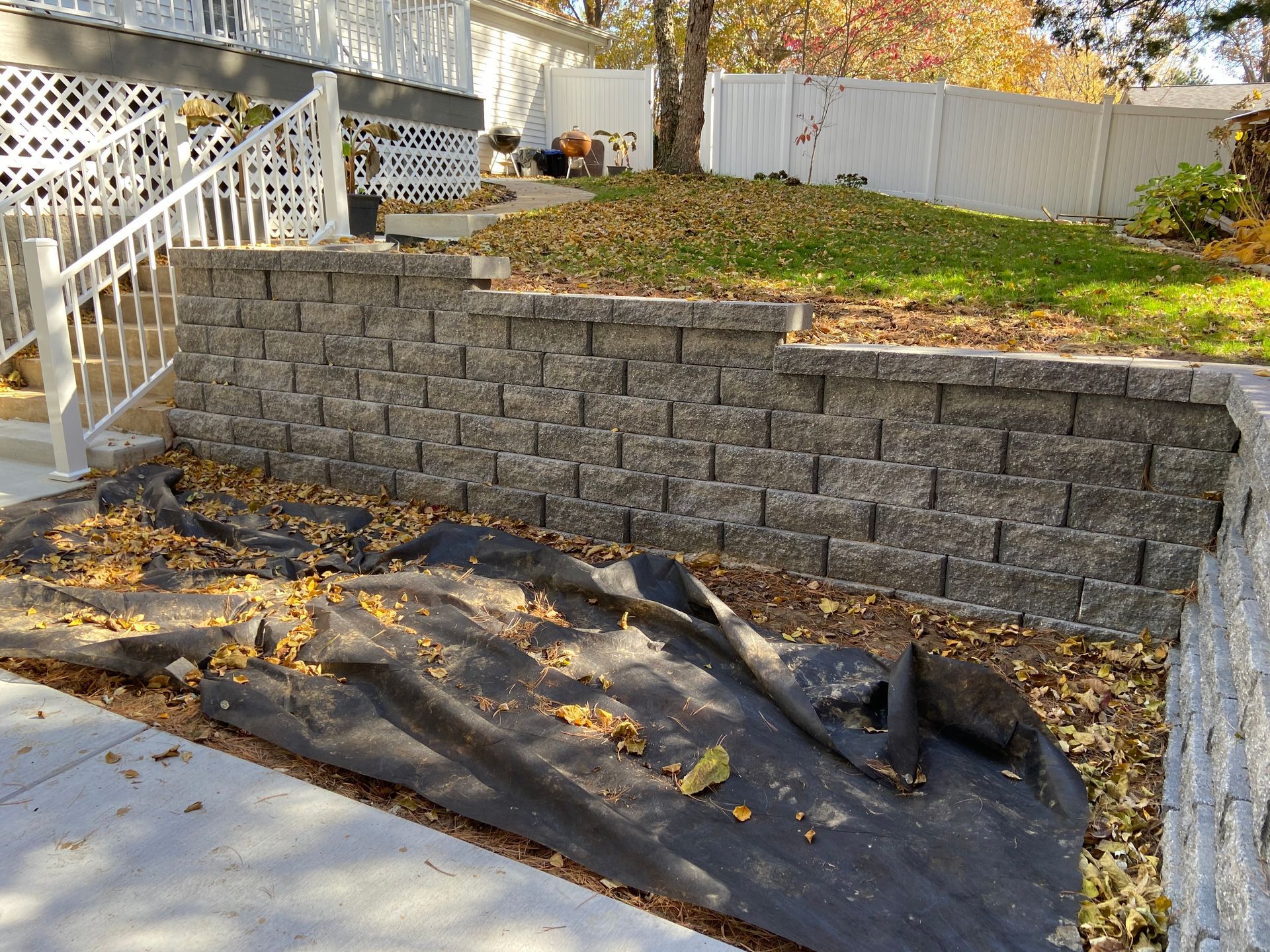 Retaining wall made of gray blocks. Black landscaping fabric covered in leaves on the ground.