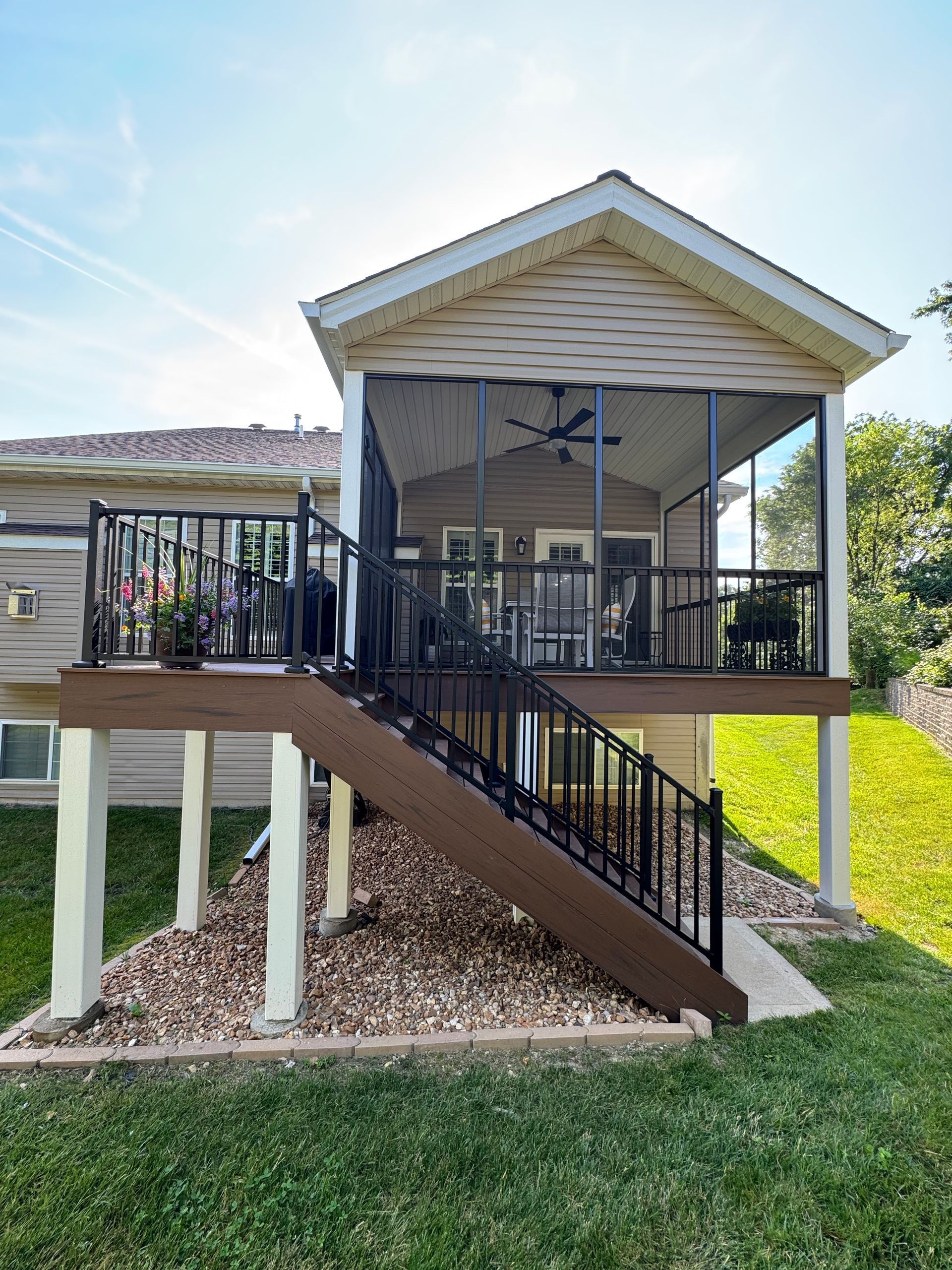 Covered wooden deck with outdoor dining table, grill, and black railing overlooking a neighborhood.