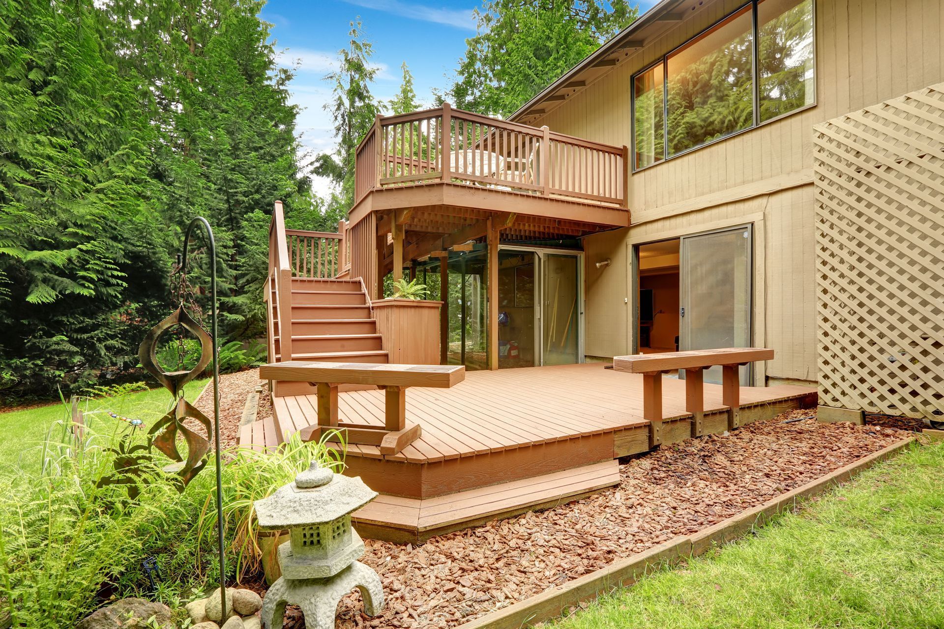 Two-story house with wooden decks, stairs, and a garden. Beige exterior, brown decks. Lush green trees.