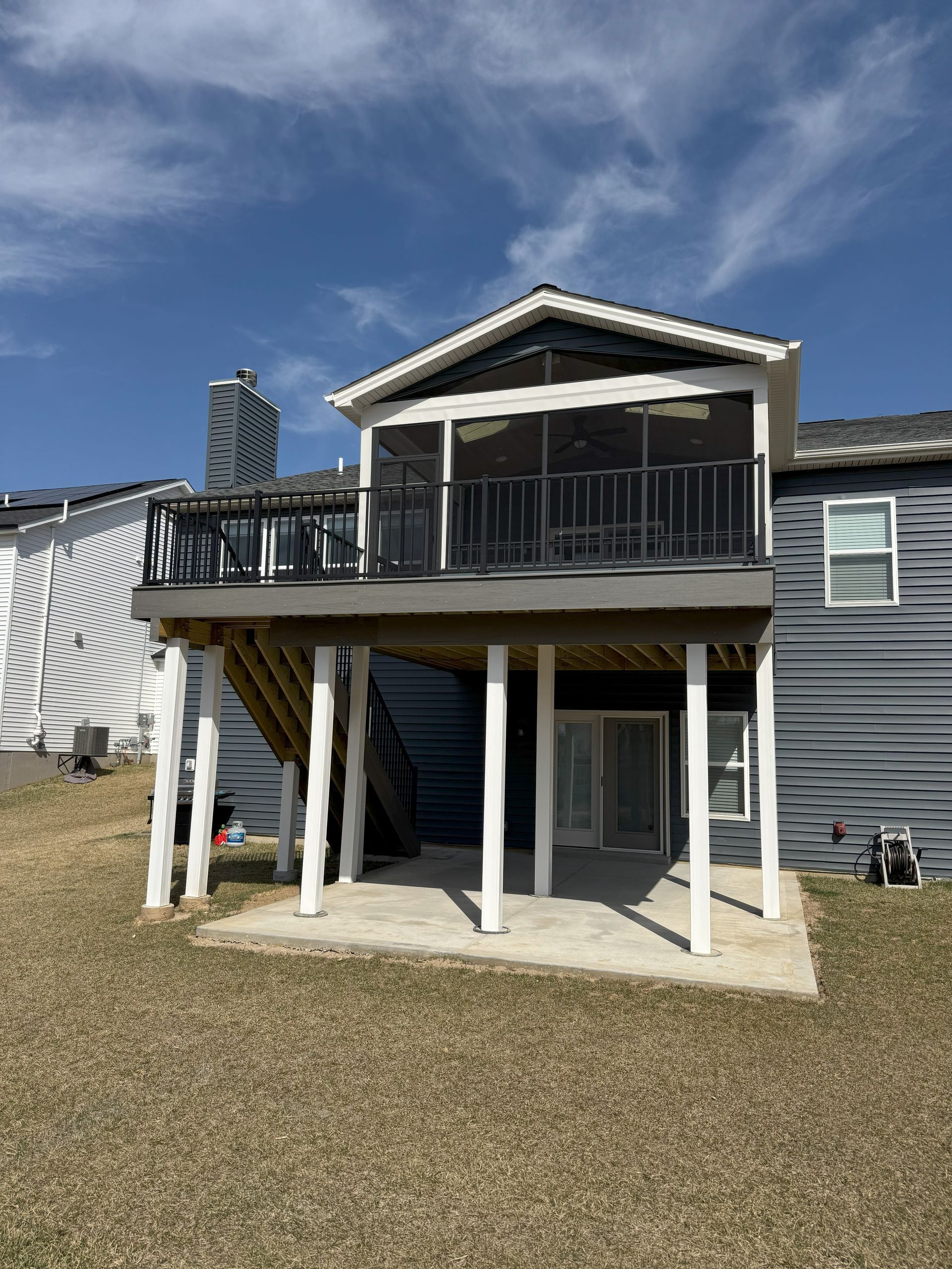 Screened-in deck with white supports, grey siding, and stairs. Blue sky and grassy yard in the background.