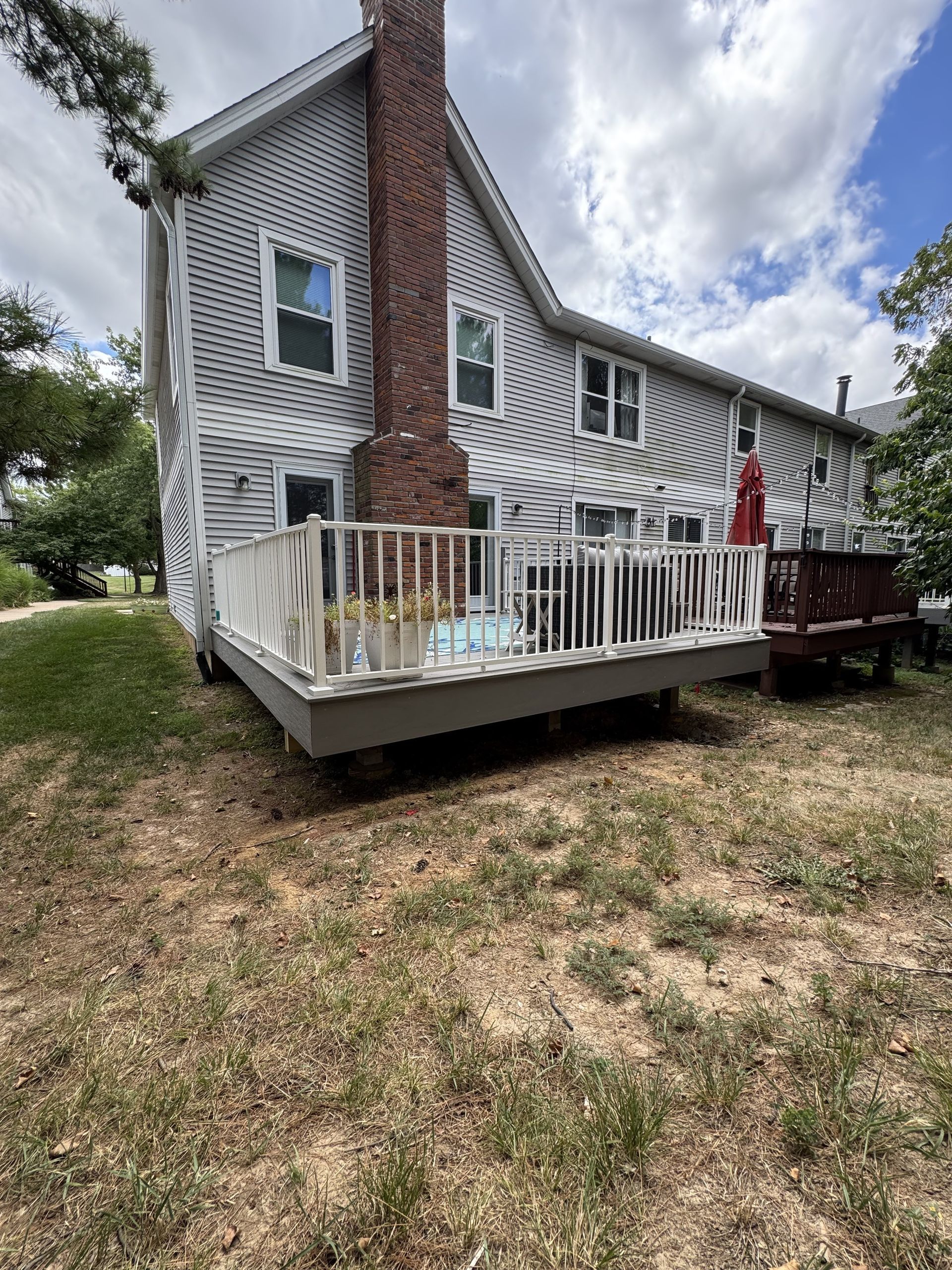 Back of a gray house with a deck. Red brick chimney, white railing, and brown yard.