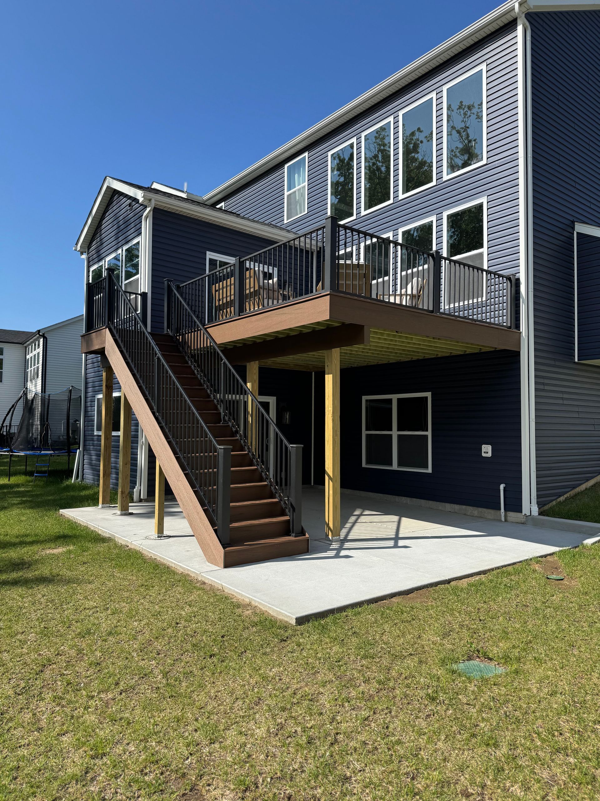 Back of a two-story blue house with a wooden deck and staircase leading to a concrete patio on a sunny day.