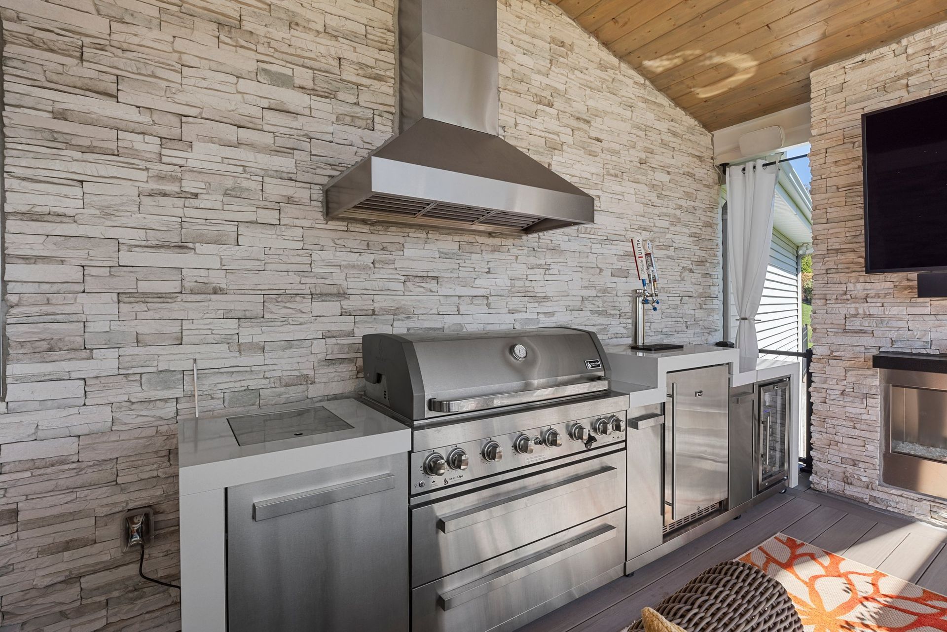 Outdoor kitchen with stainless steel grill, hood, and refrigerator on a stone wall.