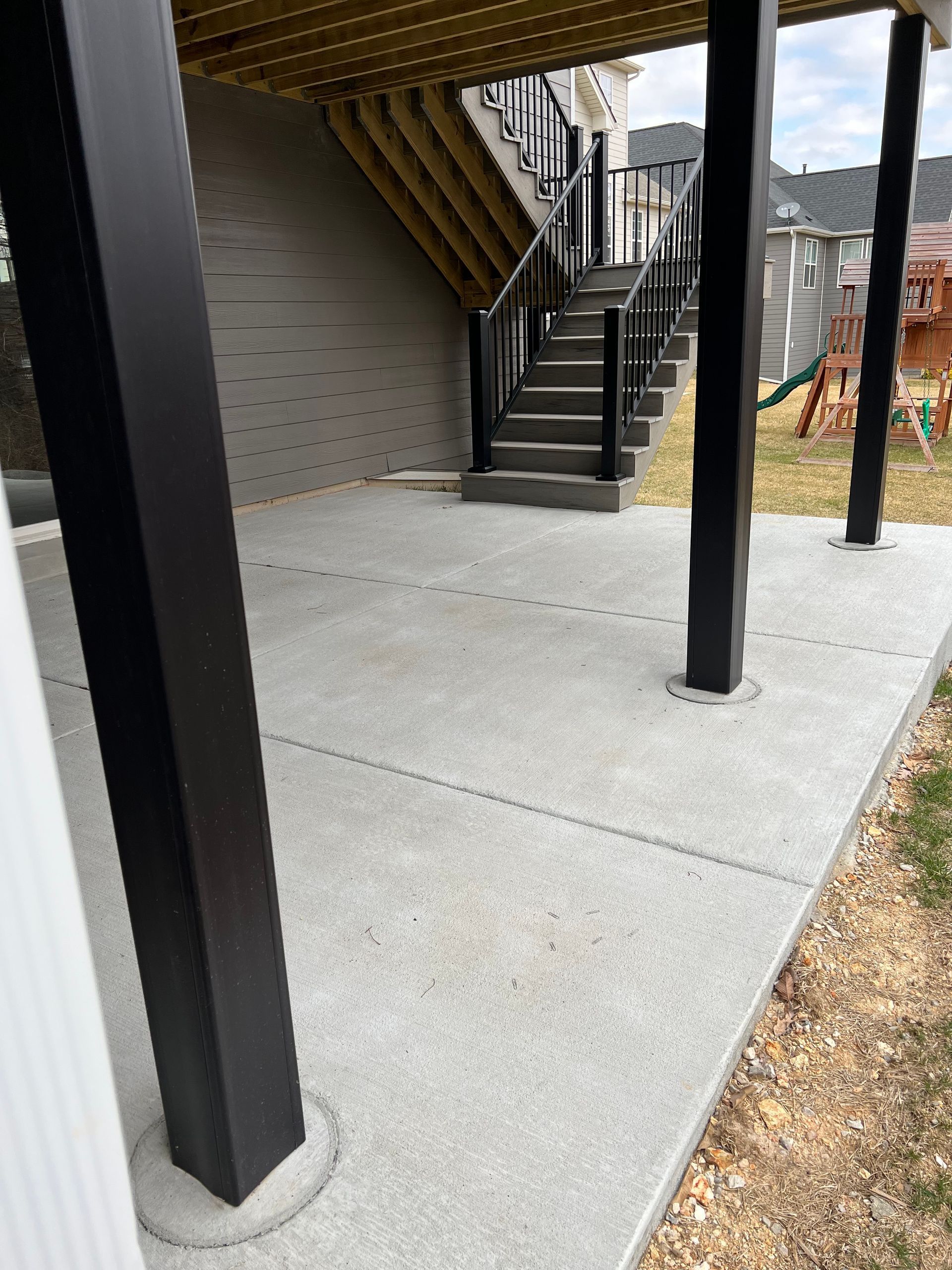 Concrete patio under a deck with black support beams and stairs leading up.