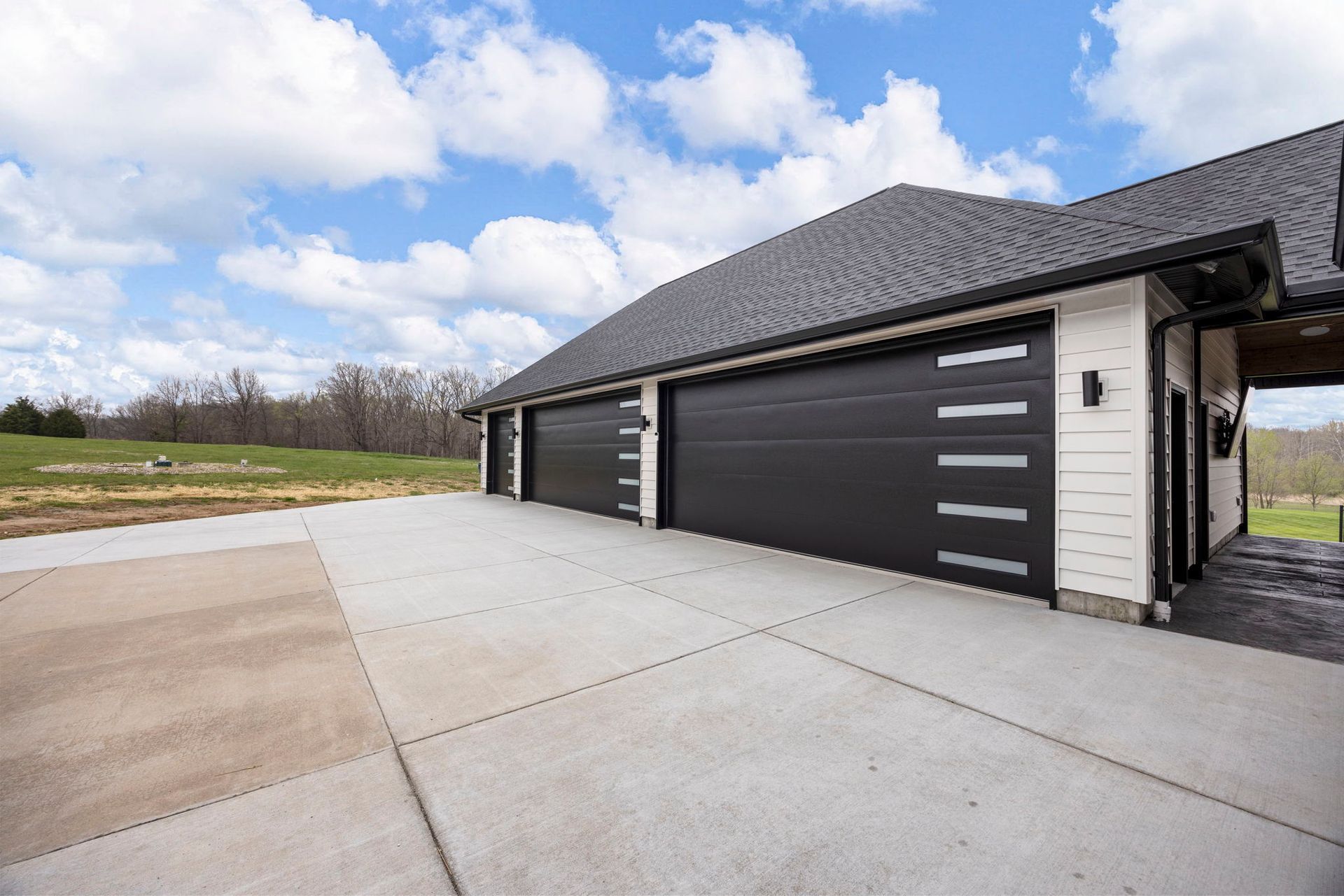 Three-car garage with black doors and concrete driveway under a cloudy blue sky.