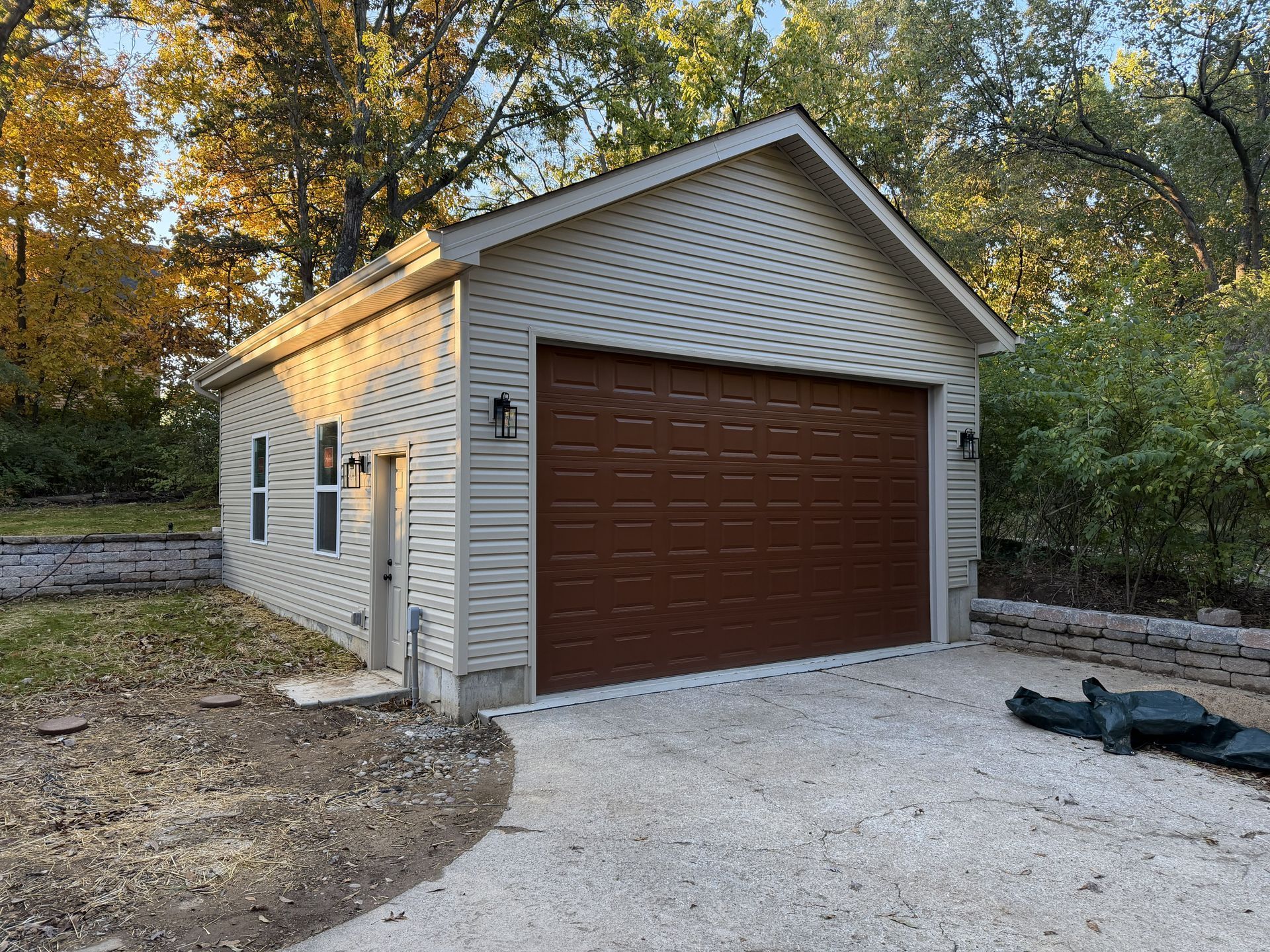 Tan garage with brown door, side entrance, and gravel driveway. Set in a wooded area.