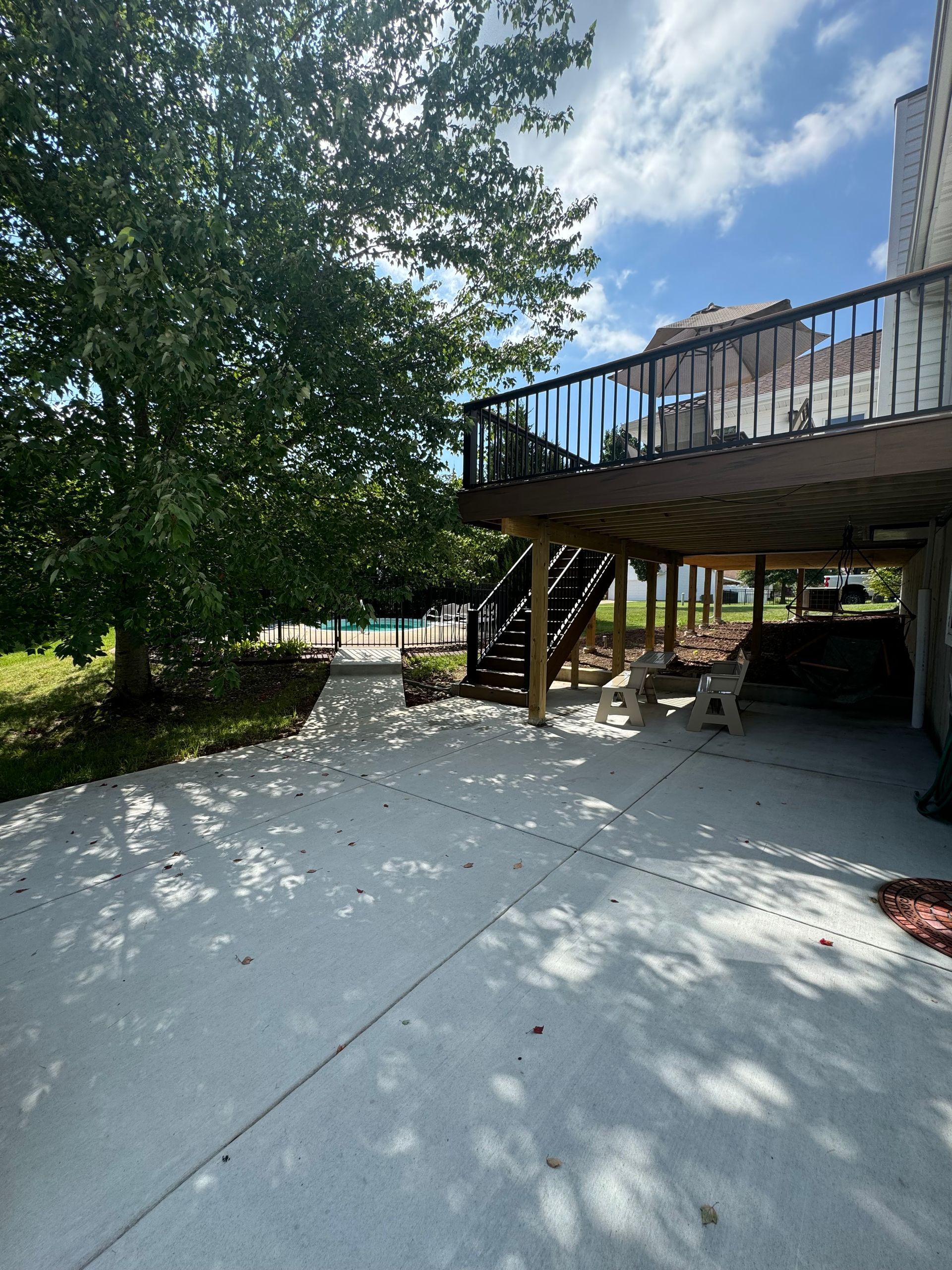 A backyard with a concrete patio, wooden deck with stairs, a tree, and a glimpse of a pool.