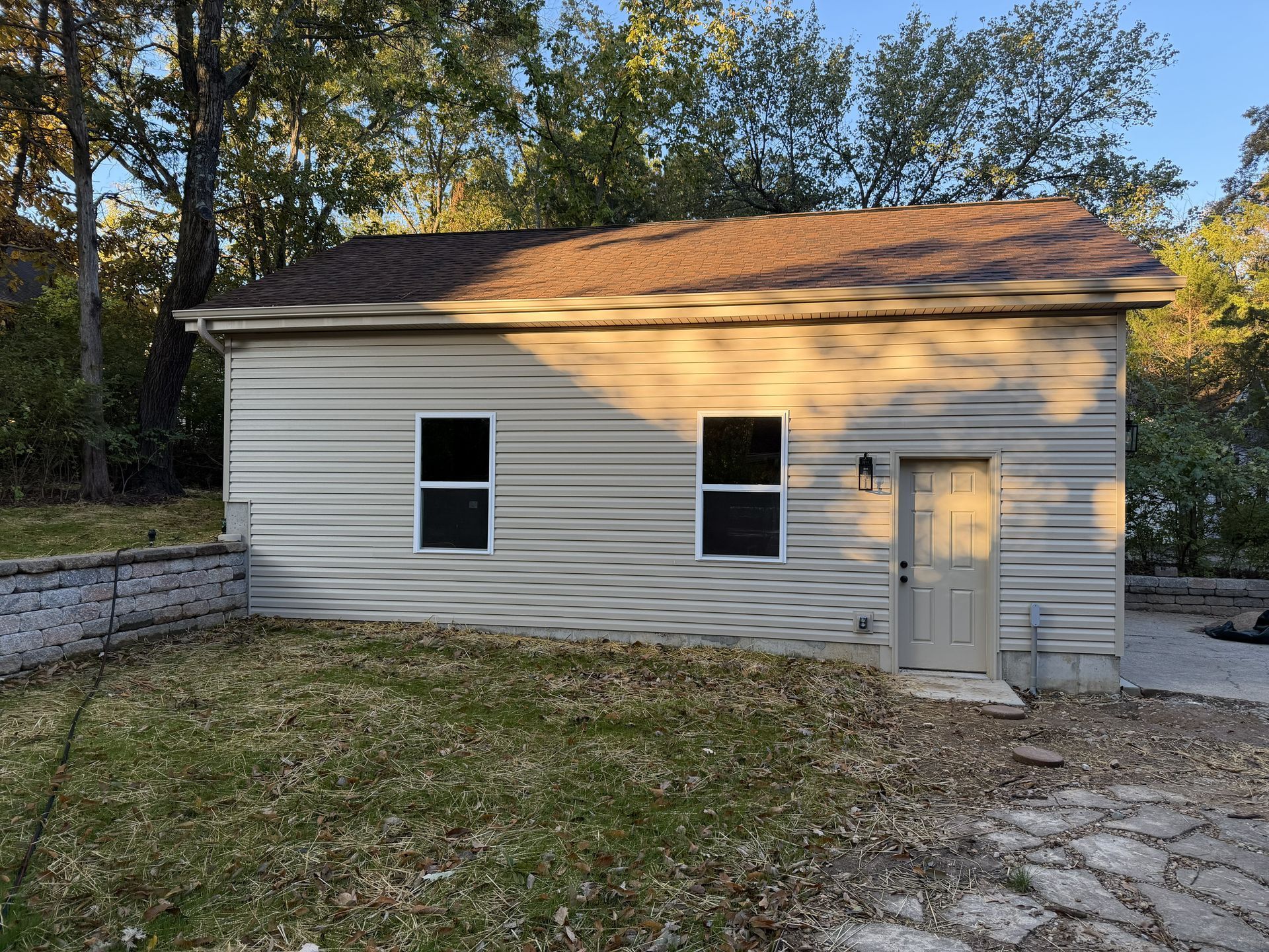 Tan building with two windows and a door, brown roof, set against trees.