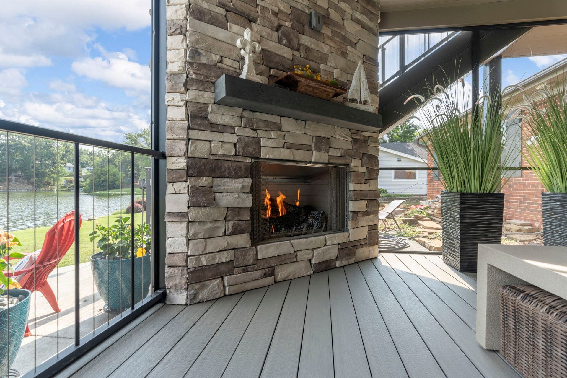Outdoor deck with stone fireplace, fire burning, lake view, black railing, and potted plants.