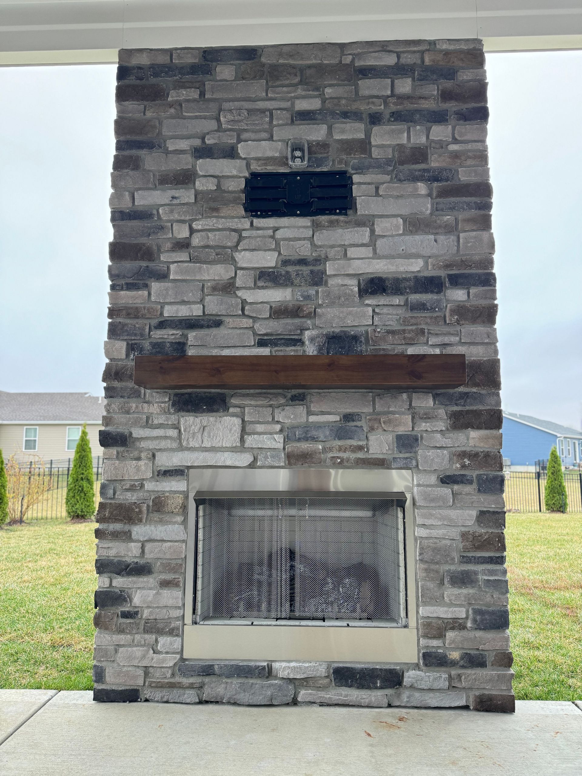 Outdoor stone fireplace with a stainless steel firebox, wooden mantel, and brick facade.
