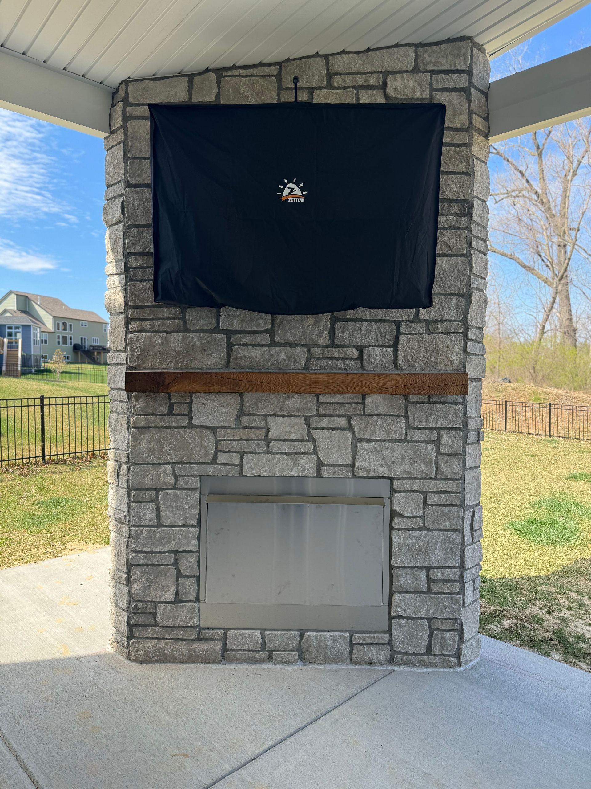 Stone fireplace with covered TV and wooden mantel on a patio.
