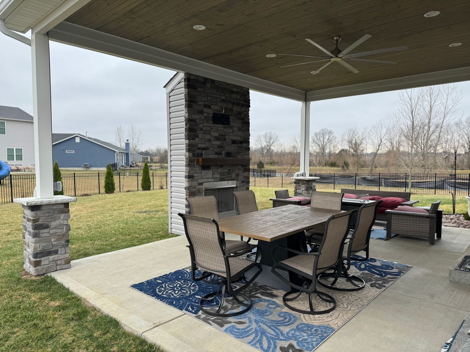 Covered patio with dining set, outdoor fireplace, and ceiling fan. Overlooking a yard and distant houses.
