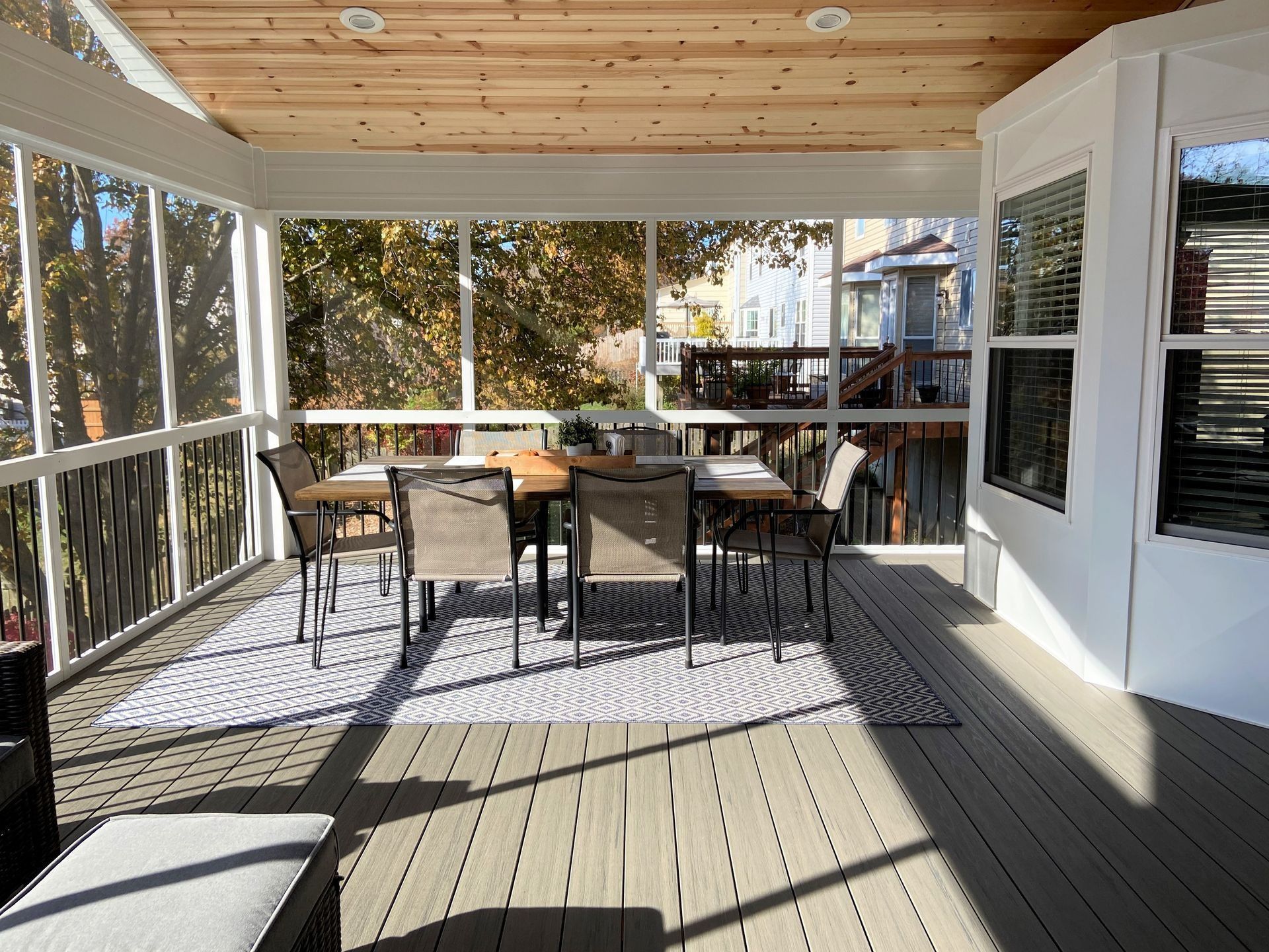 Screened-in porch with dining table and chairs, gray deck, white trim, and a view of trees and buildings.