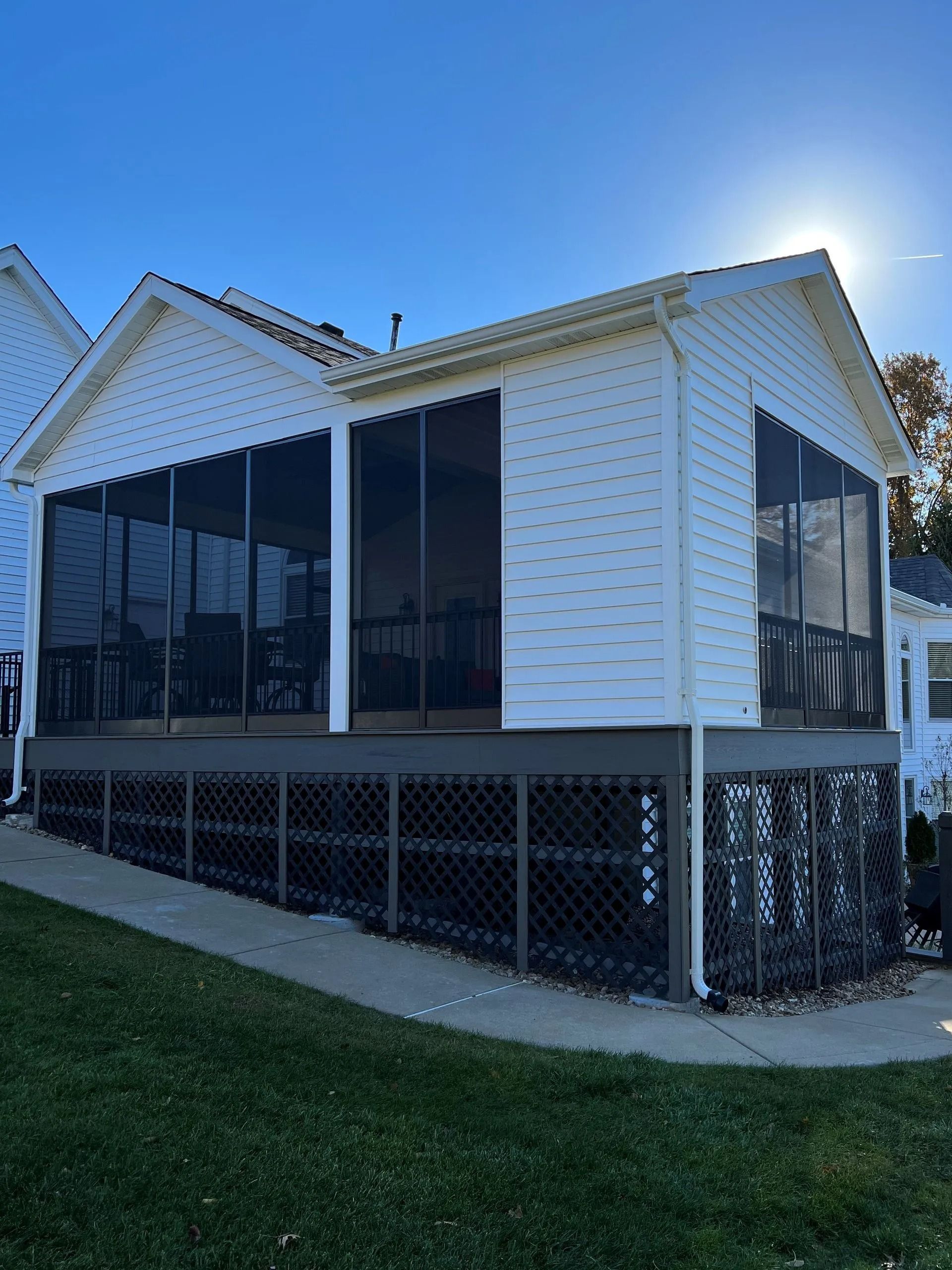 Screened-in porch on a raised deck, white siding, brown lattice and railing, dark screening with a sunny sky.