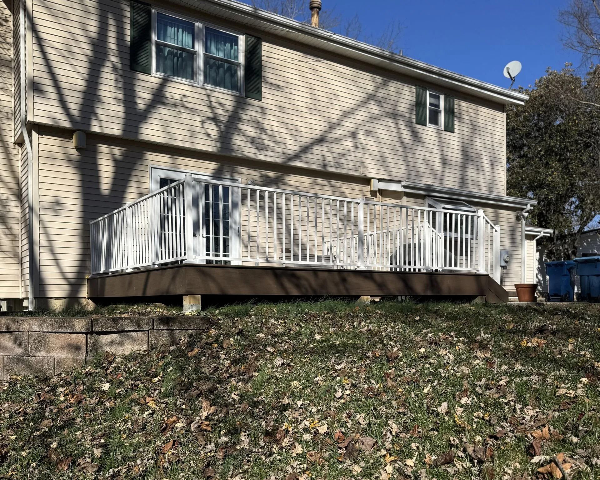 Back of a two-story beige house with a brown wooden deck and white railings. Green grass and fall leaves in the yard.