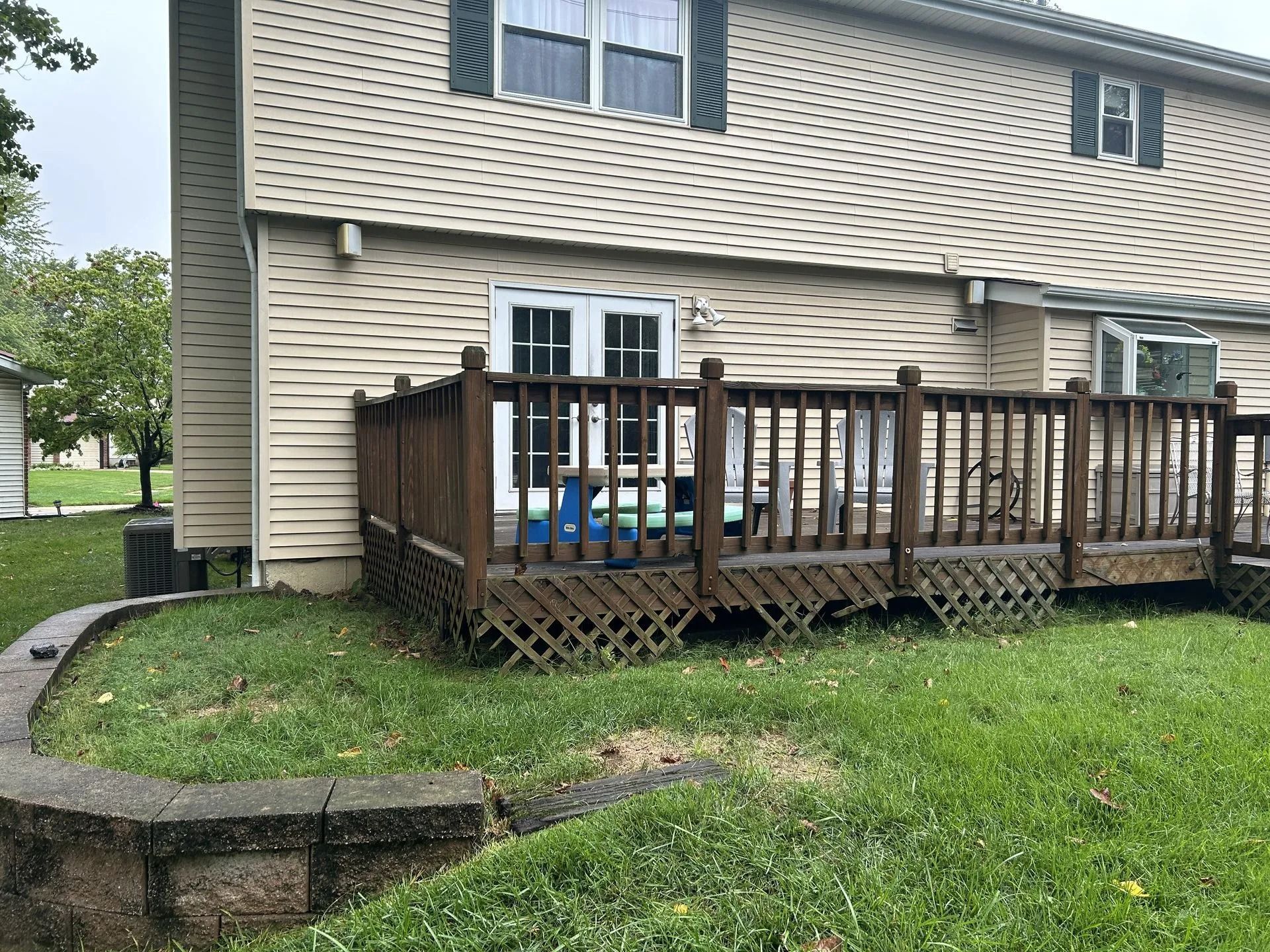 Back of a two-story beige house with a brown wooden deck and a small retaining wall in a grassy yard.