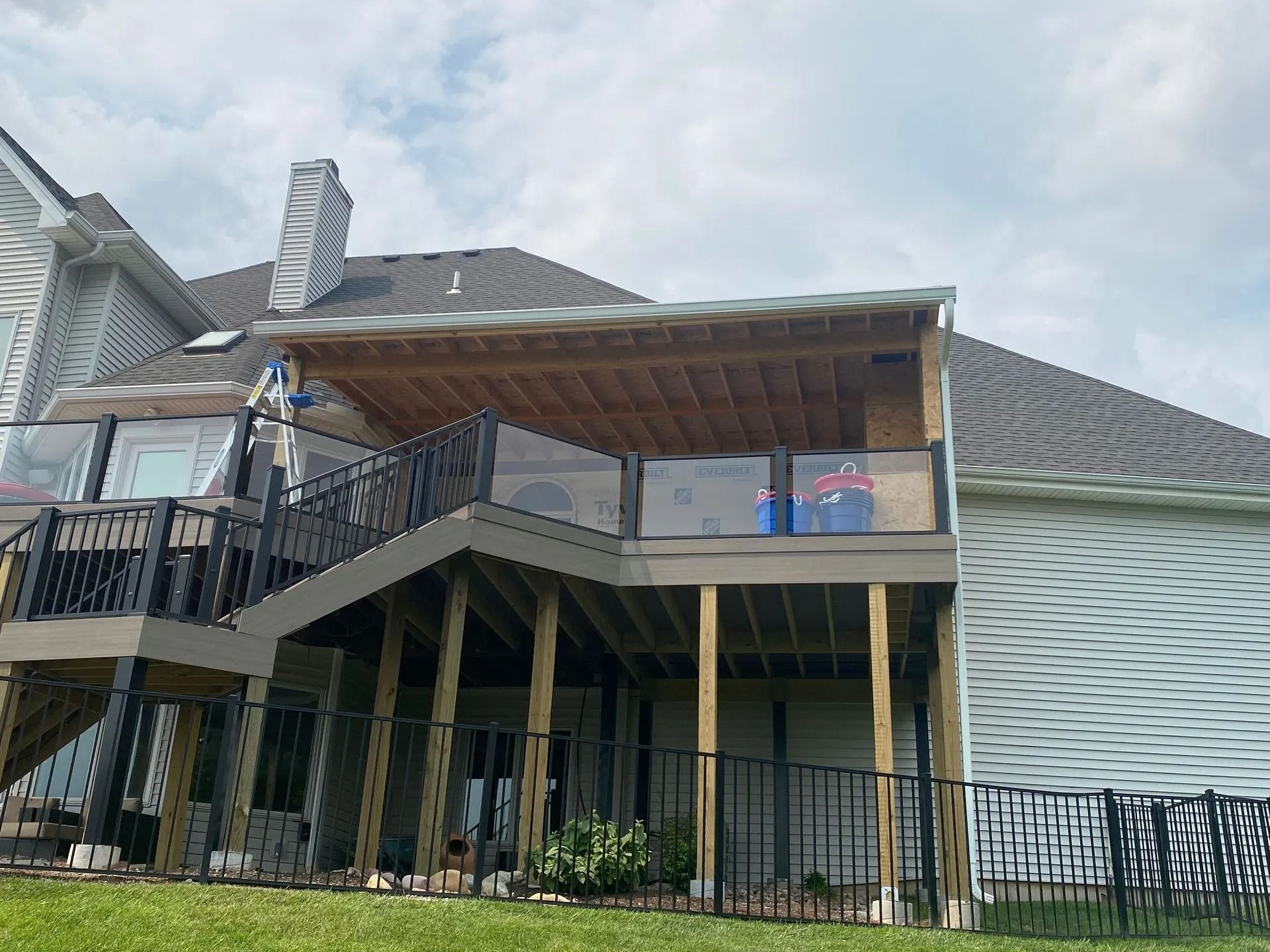 Two-story wooden deck attached to a house with black railings, clear glass panels, and a covered upper deck.