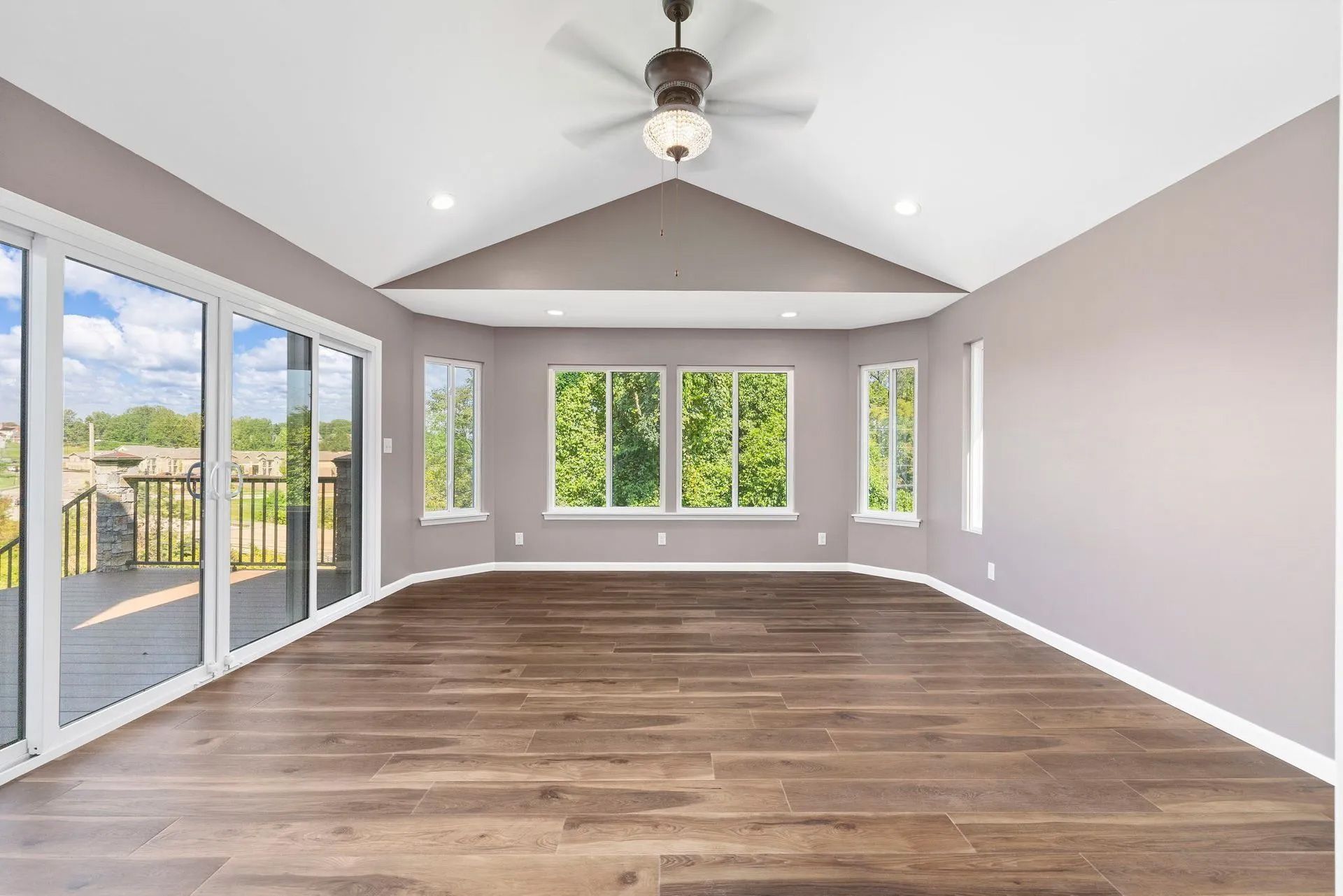 Empty room with wooden floors, large windows, and a ceiling fan; painted in neutral tones.