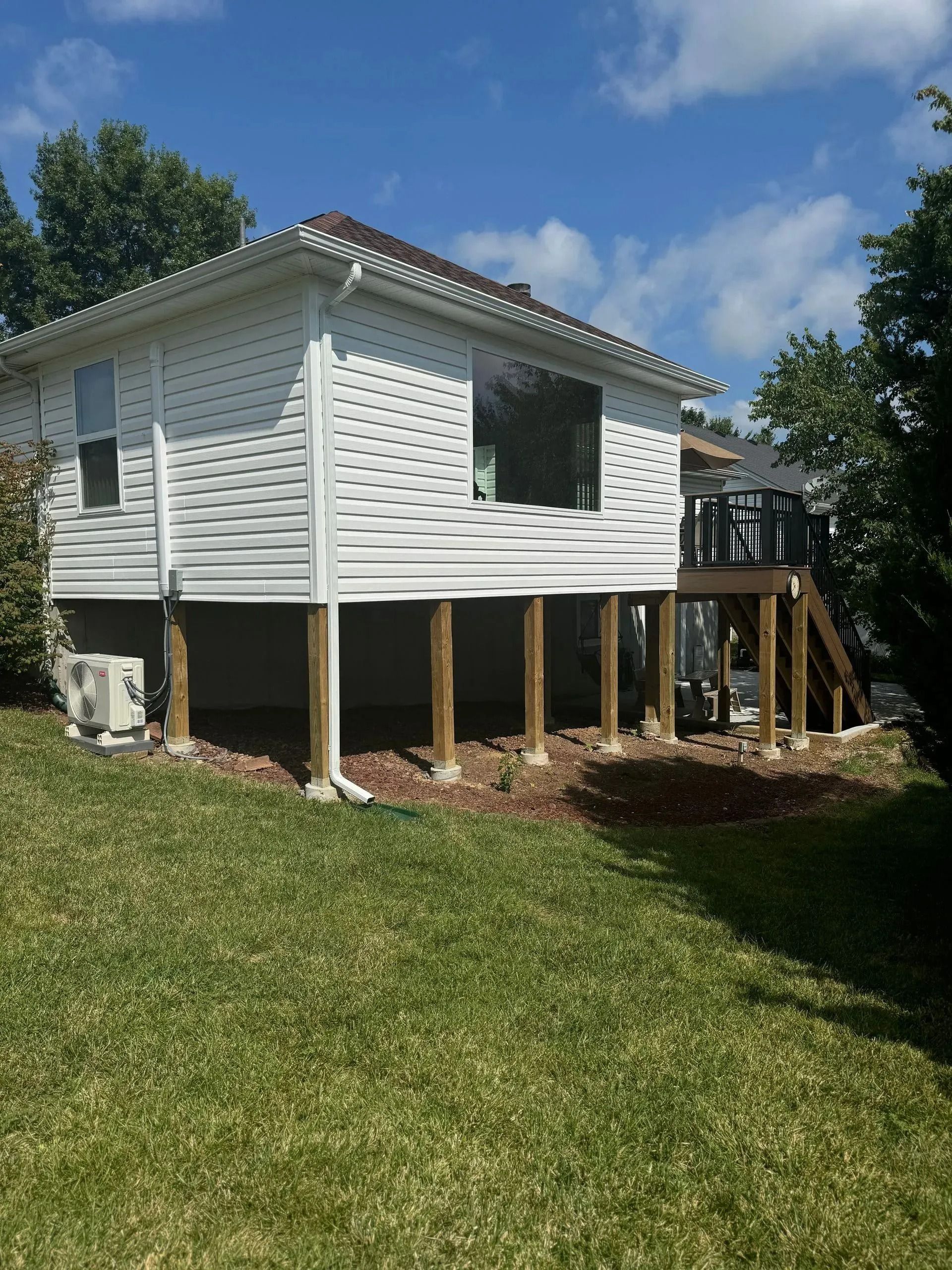 House with white siding and brown deck supports, set on a green grassy hill under a blue sky.