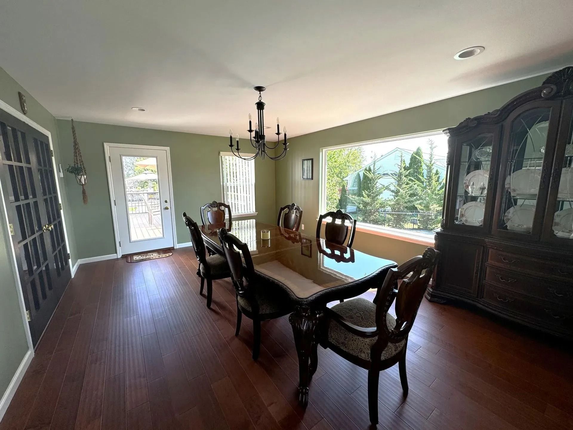 Dining room with a long table, chairs, hutch, and large window. Green walls, dark wood floor.