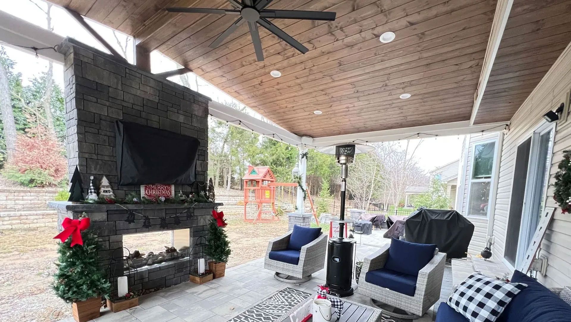 Patio with fireplace, seating, and a ceiling fan. Christmas decorations. Outdoor setting.