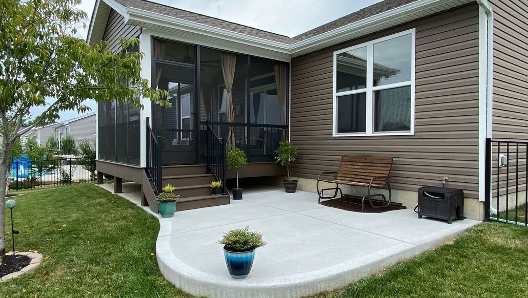 Screened-in porch and concrete patio adjoin a brown house with a yard.  A bench and potted plants are present.