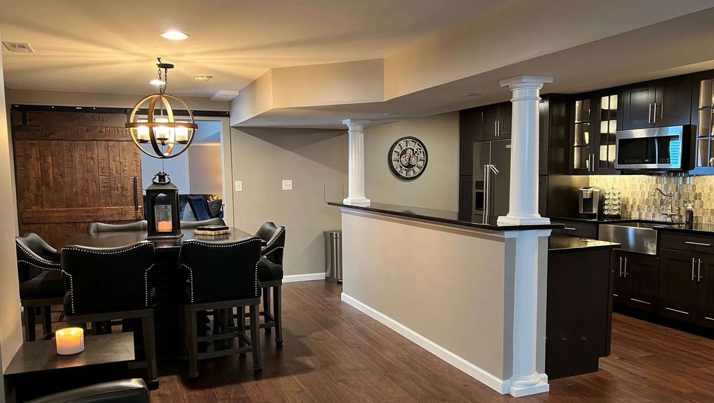 A finished basement with a dining area, kitchen, and bar. Dark wood cabinets, black countertops, and dim lighting.