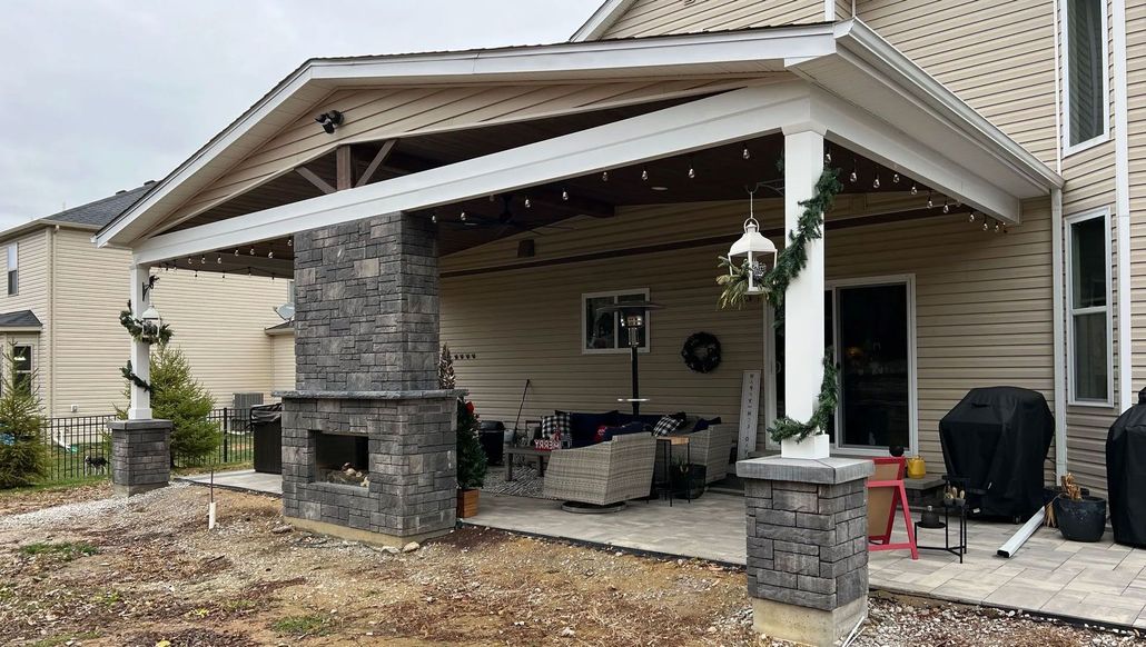 Covered patio with fireplace, seating area, and decorative lights. Beige siding, gray stone accents.