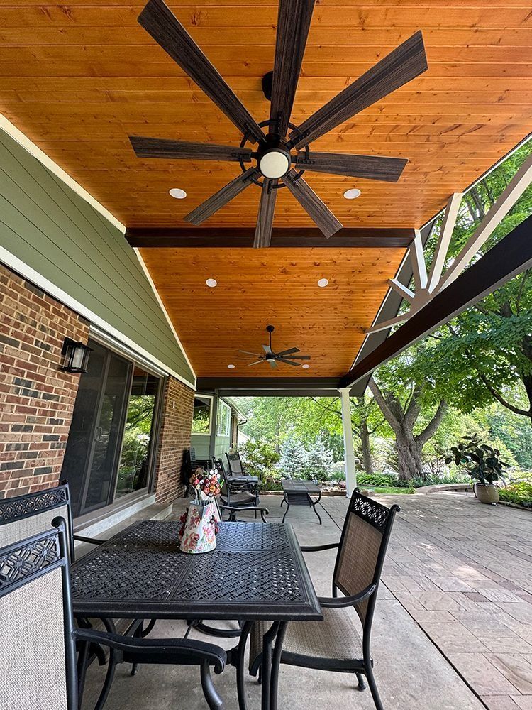 Patio with wooden ceiling, ceiling fans, and outdoor furniture. Brick wall on left, trees in background.