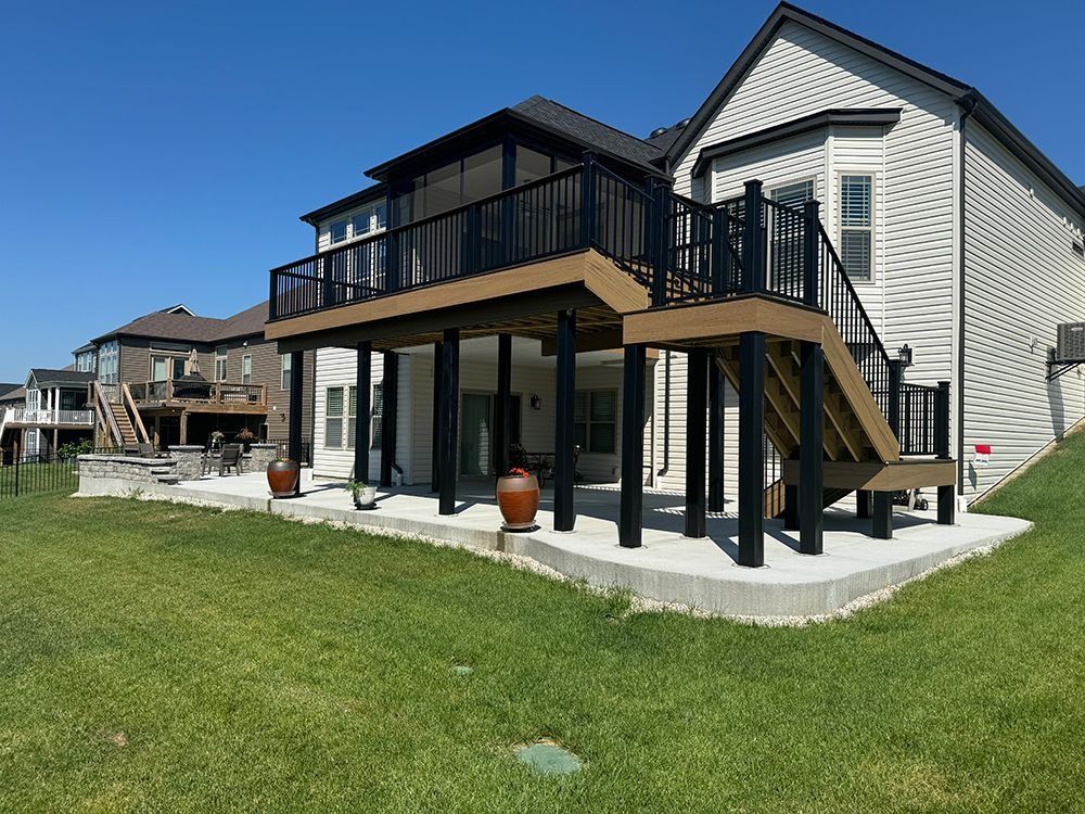 A two-story deck with black railings and brown decking, built onto a light-colored house. Green grass in foreground.