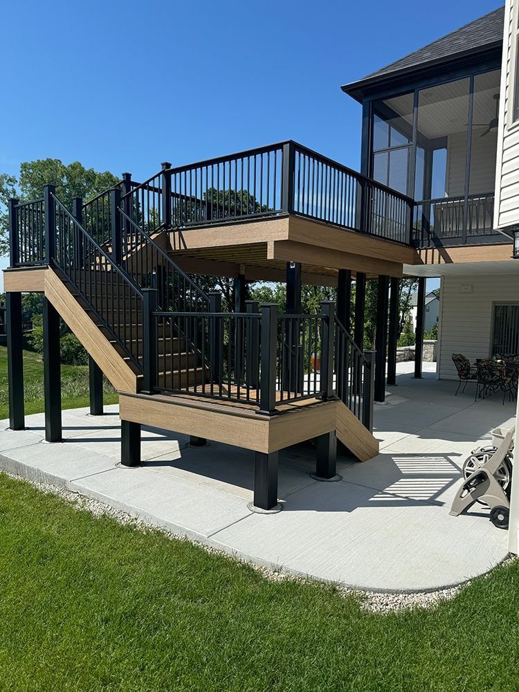 Elevated deck with stairs, black railings, attached to a house with a screened porch. Sunny outdoor setting.