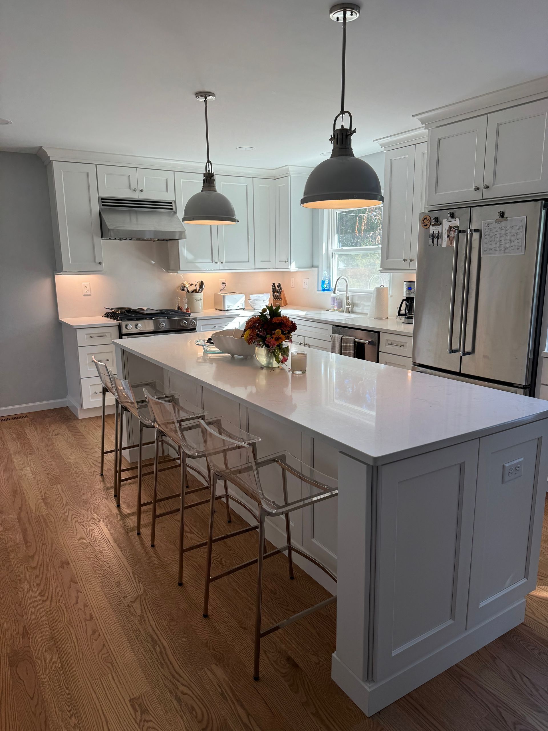 Kitchen with white cabinets, island, wooden floor, gray pendant lights, clear bar stools, and stainless steel appliances.