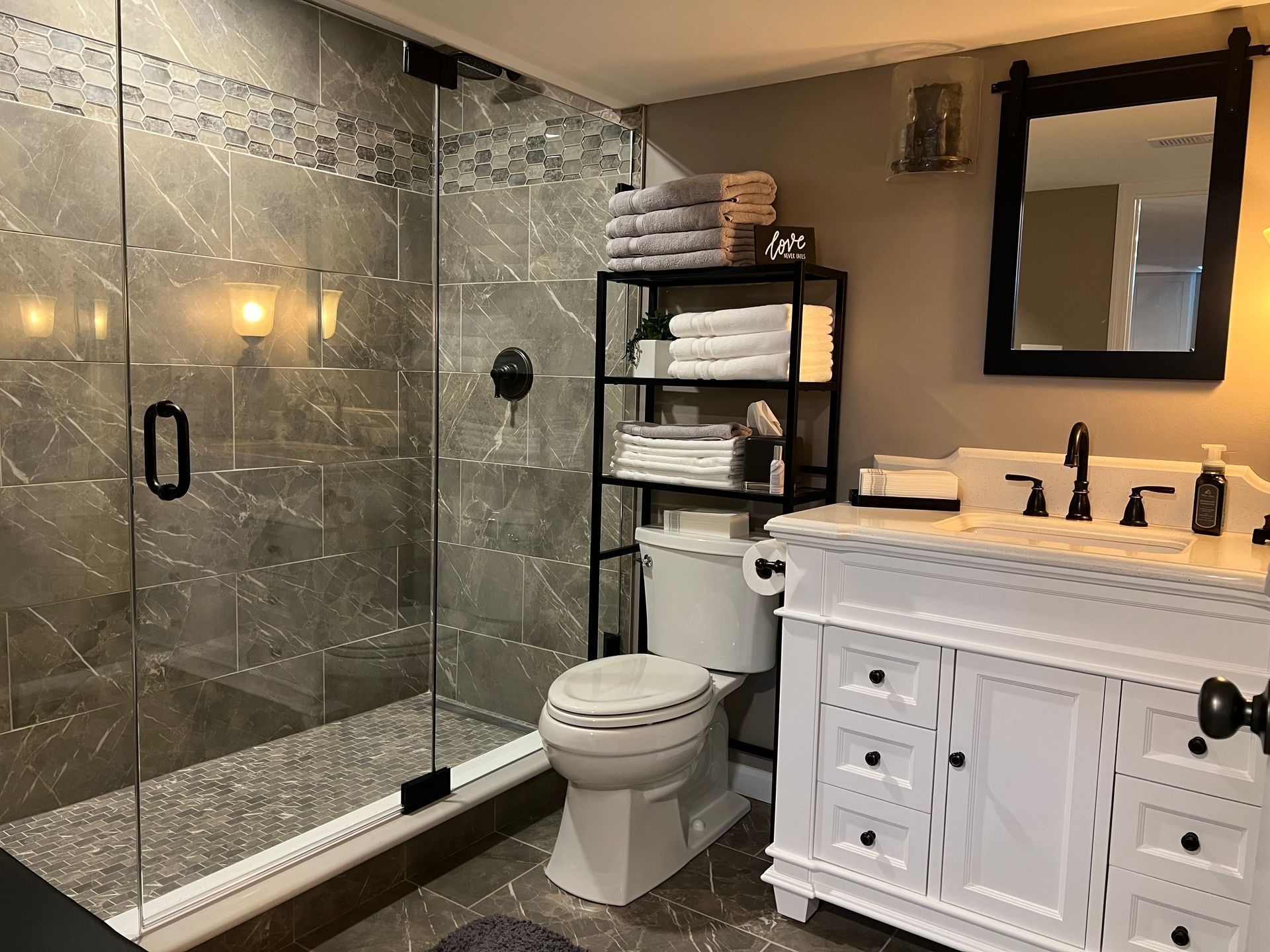 Bathroom with gray tile shower, white vanity, black shelving with towels, and a mirror.