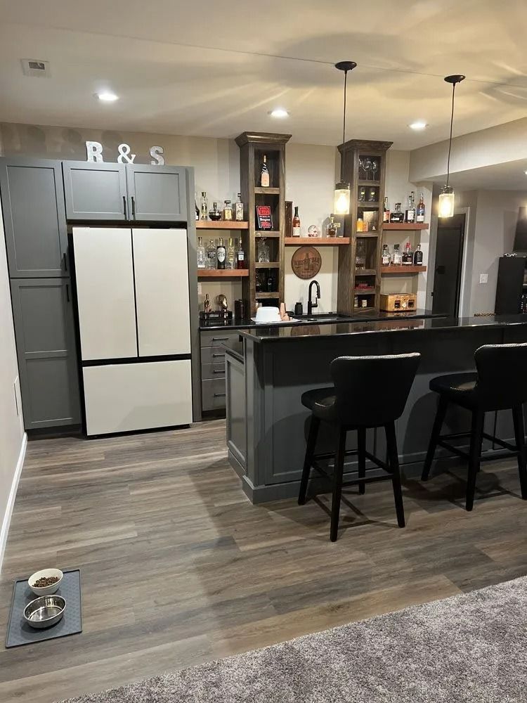 Finished basement bar with grey cabinets, refrigerator, shelving, and stools.