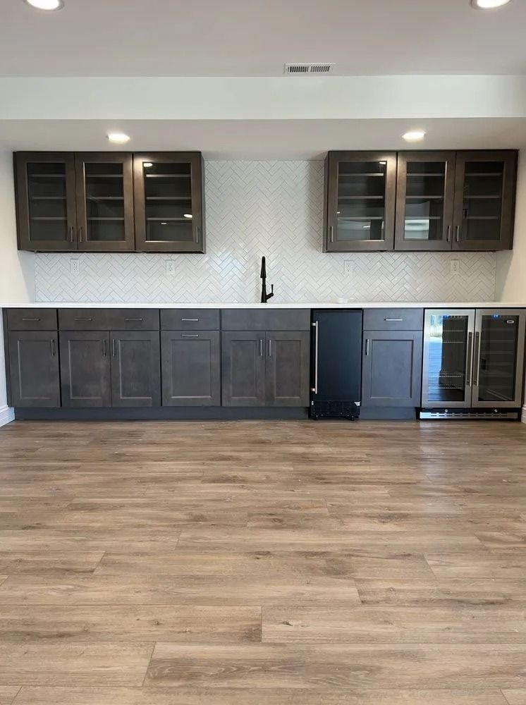 Bar area with dark gray cabinets, white backsplash, and wood-look flooring. Includes a sink and two beverage coolers.